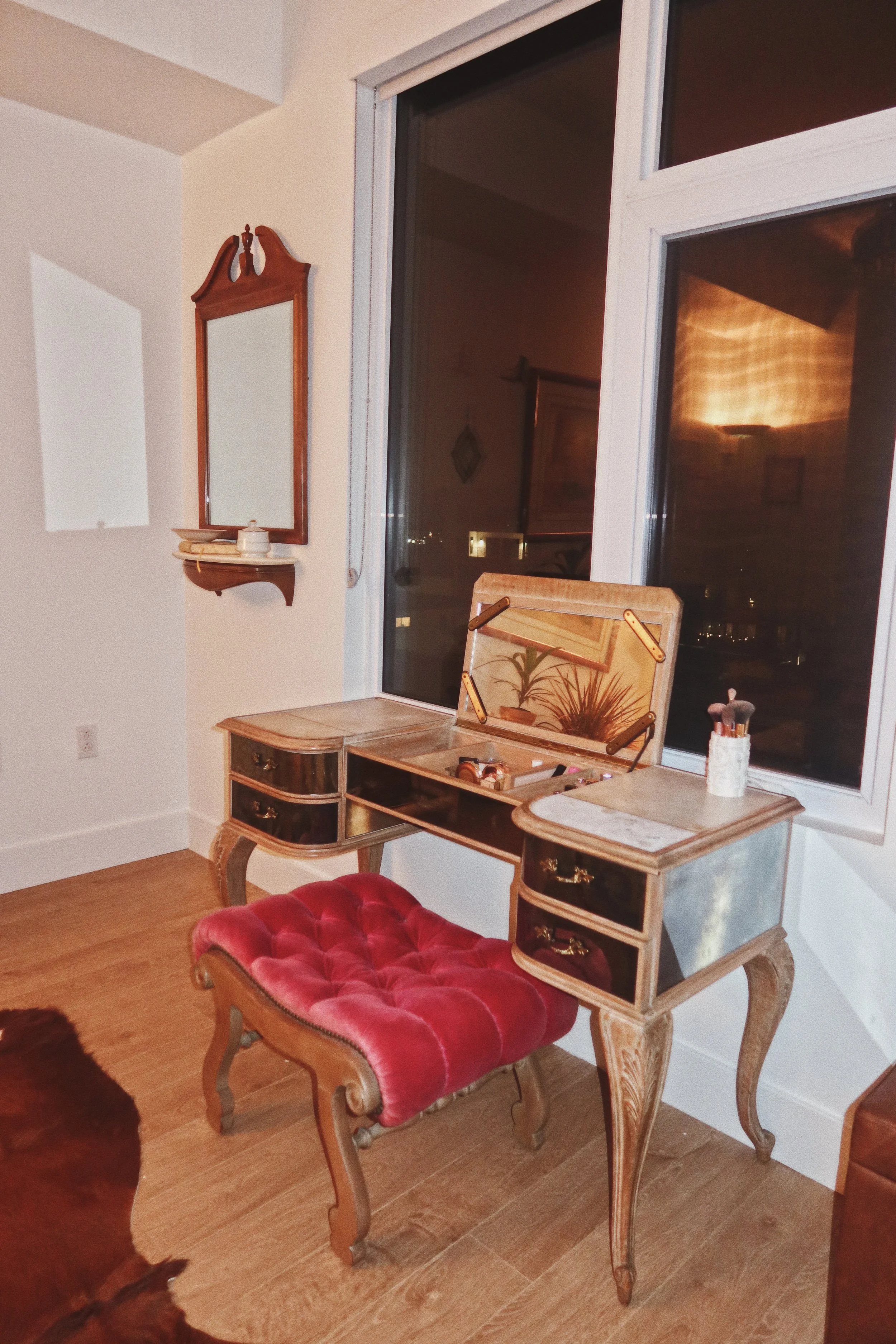 A vintage wooden vanity table with a glass top, a floral design on the back panel, and a red velvet cushioned stool. The table has three drawers with brass handles. Behind, a large window shows a night view outside, and a small wooden shelf with a ce