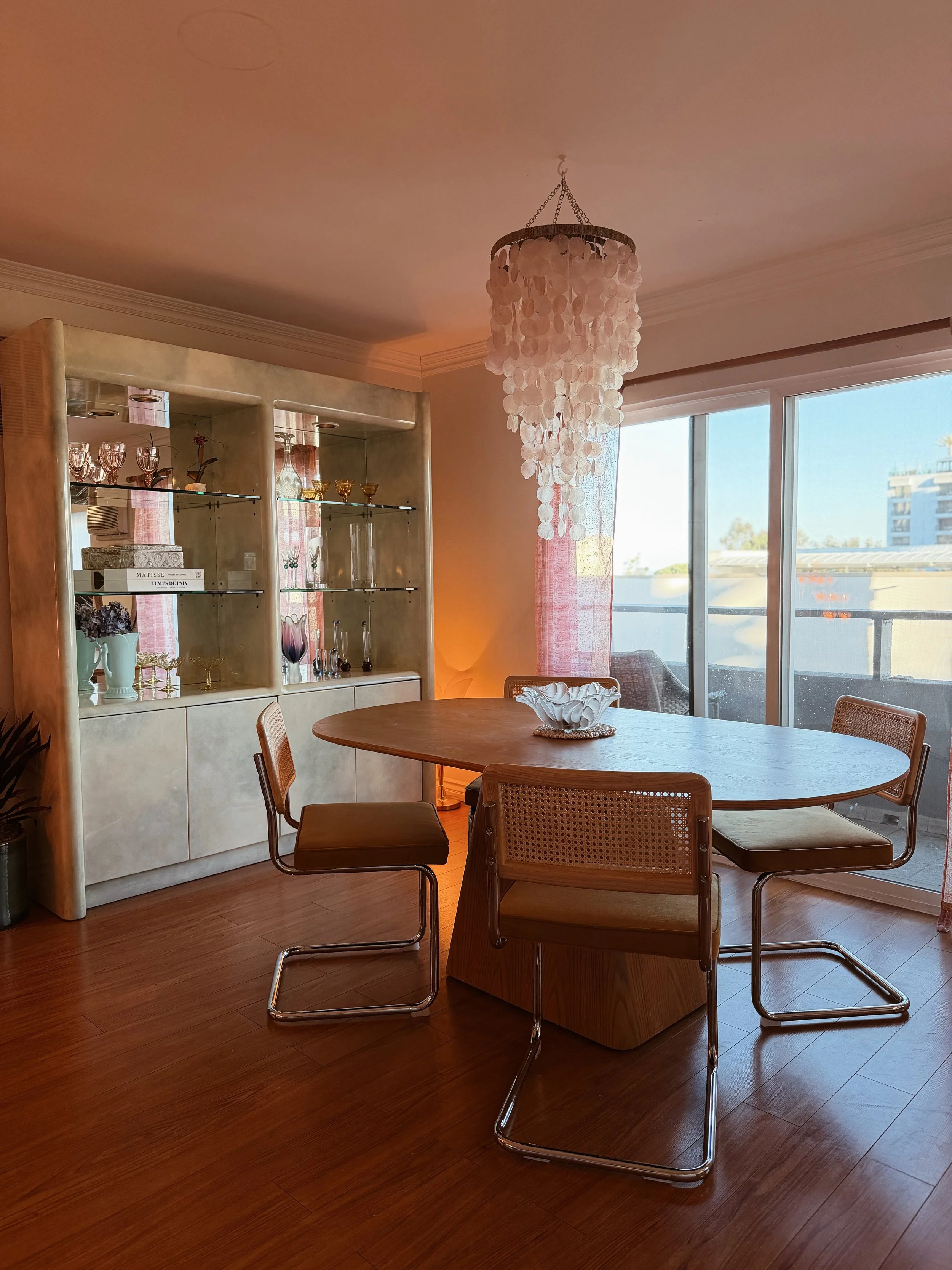 A dining room with a wooden table and four chairs, a decorative chandelier hanging from the ceiling, large windows with pink curtains allowing natural light, and a display cabinet with glassware and decorative items.