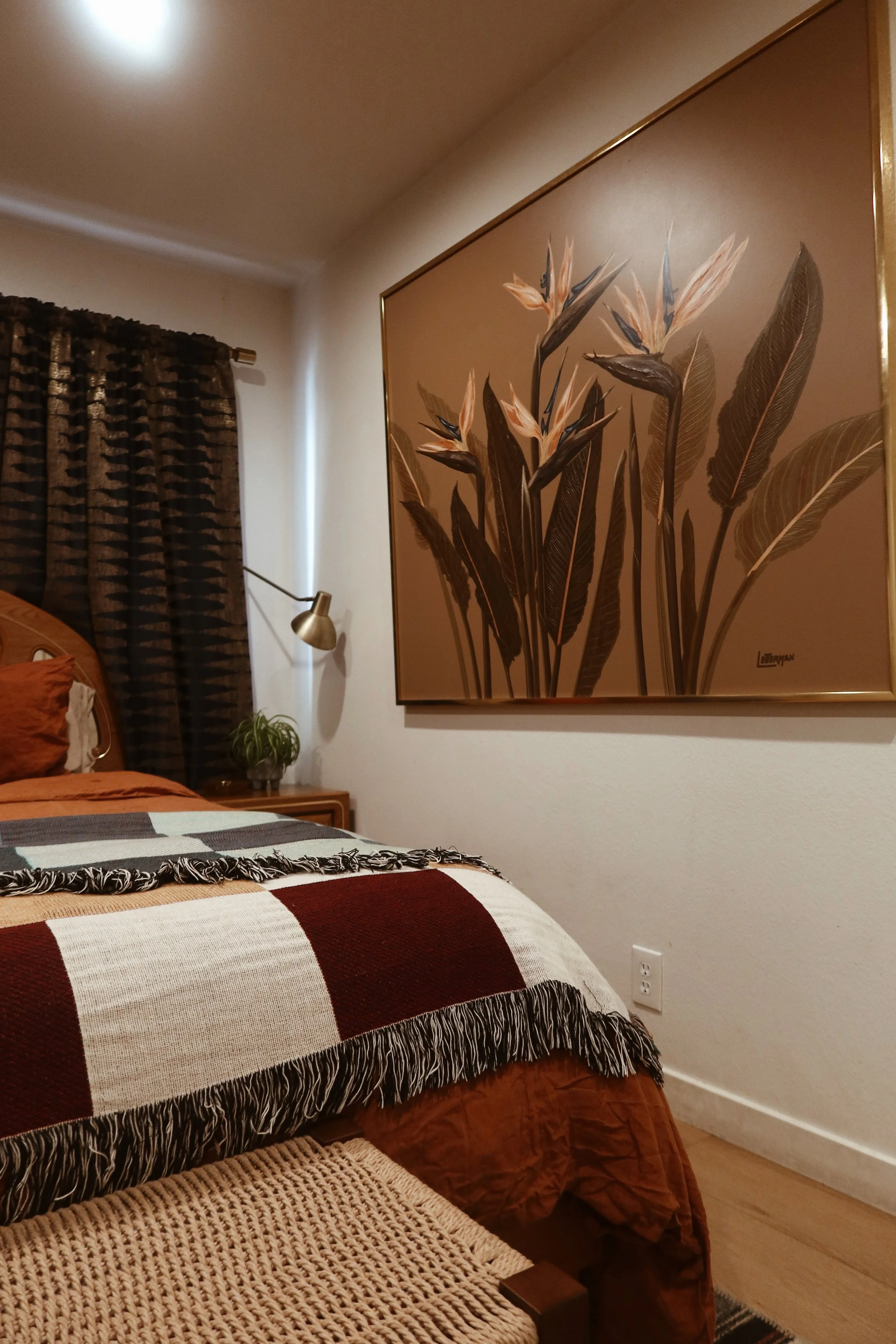 A bedroom with a bed covered by a red, beige, and black checkered blanket, a wall art of large leaves and flowers, a small bedside table with a potted plant, a brass wall lamp, and curtains with a geometric pattern.