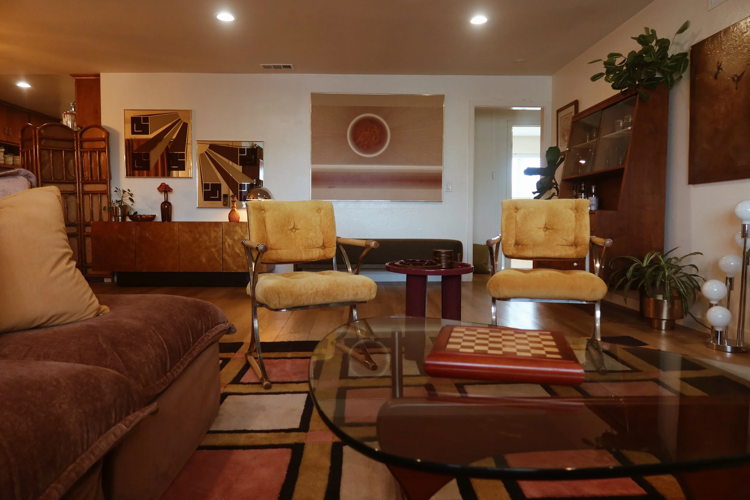A cozy living room with retro furniture including yellow armchairs and a brown sofa, decorated with plants, abstract art, and a checkerboard chessboard on a glass coffee table, with artwork on the walls and a patterned rug on the wooden floor.