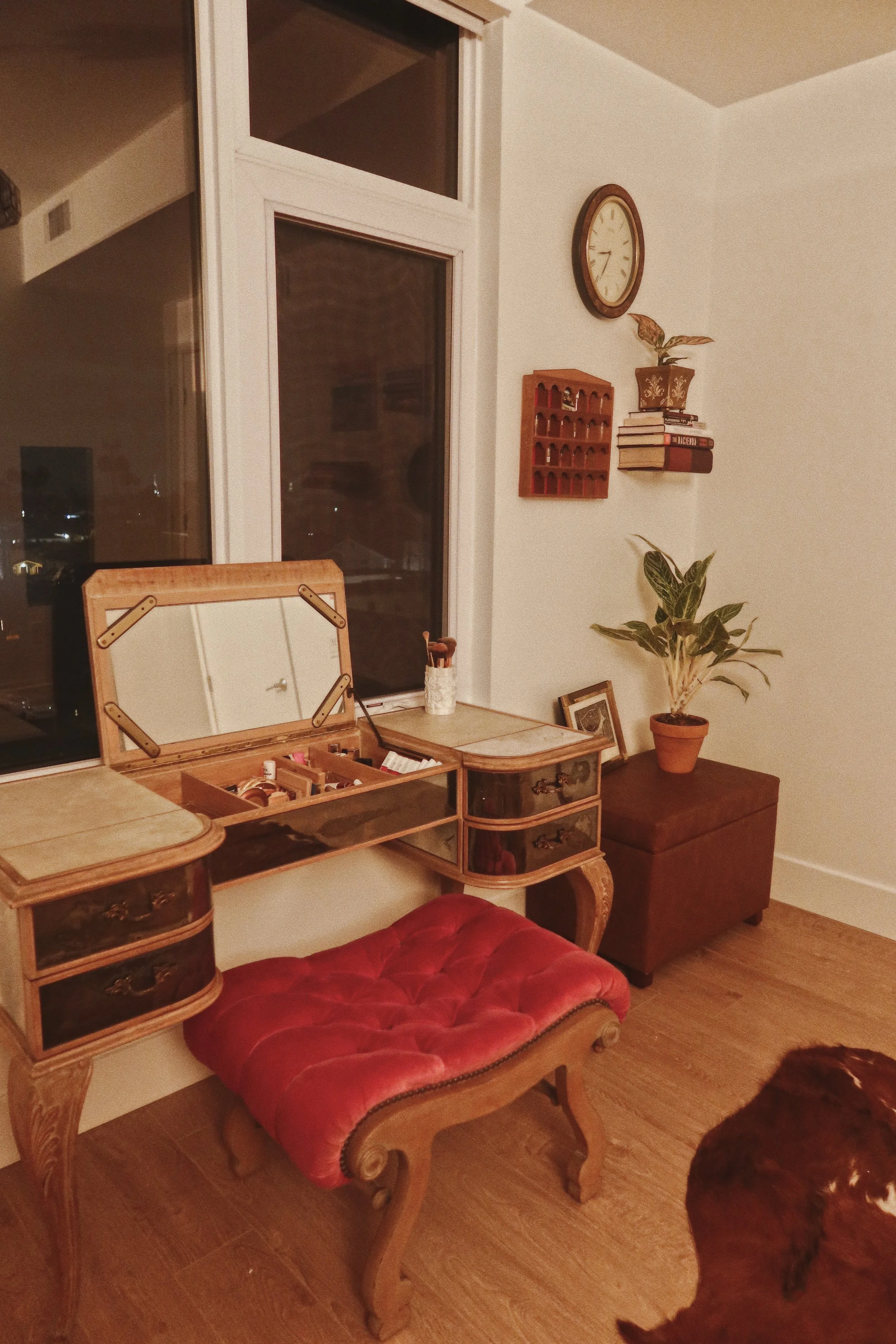 A vintage wooden vanity table with a mirror, adorned with makeup supplies, in a room with a window, potted plant, clock on the wall, and a brown dog sitting on the floor nearby.