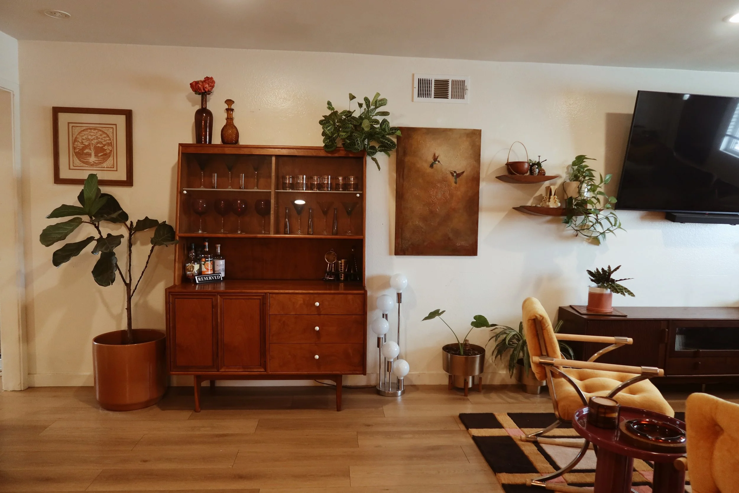 Living room with vintage wooden cabinet, houseplants, wall art, shelves, and a large flat-screen TV.