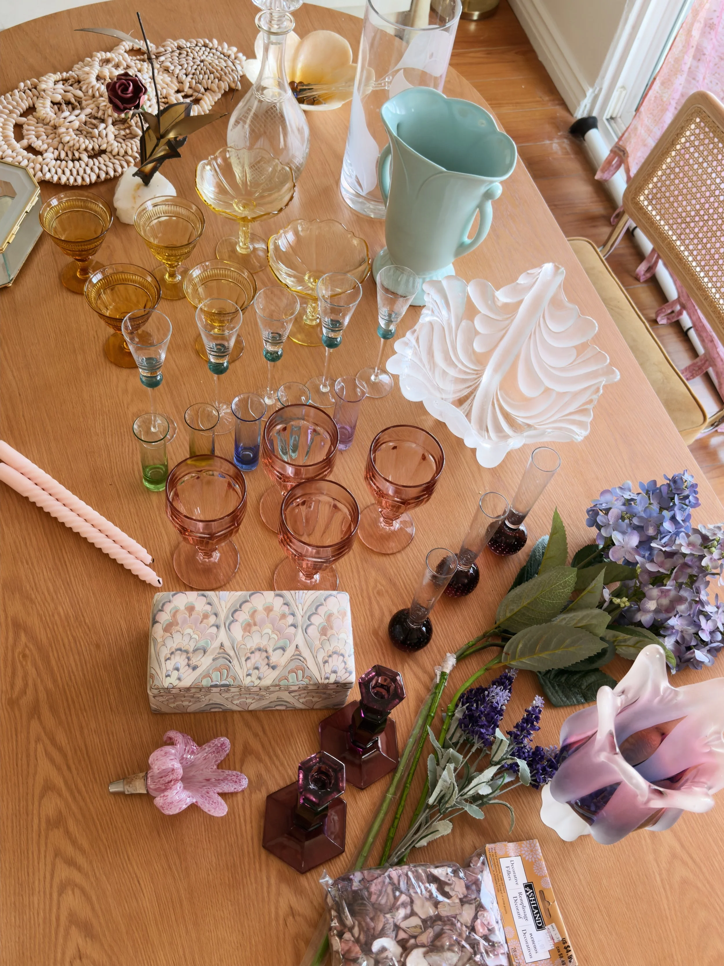 A wooden table with various decorative items including colorful glassware, vases, a ceramic jug, artificial flowers, and shells. There is also a tissue box with a marbled design and some seashells.