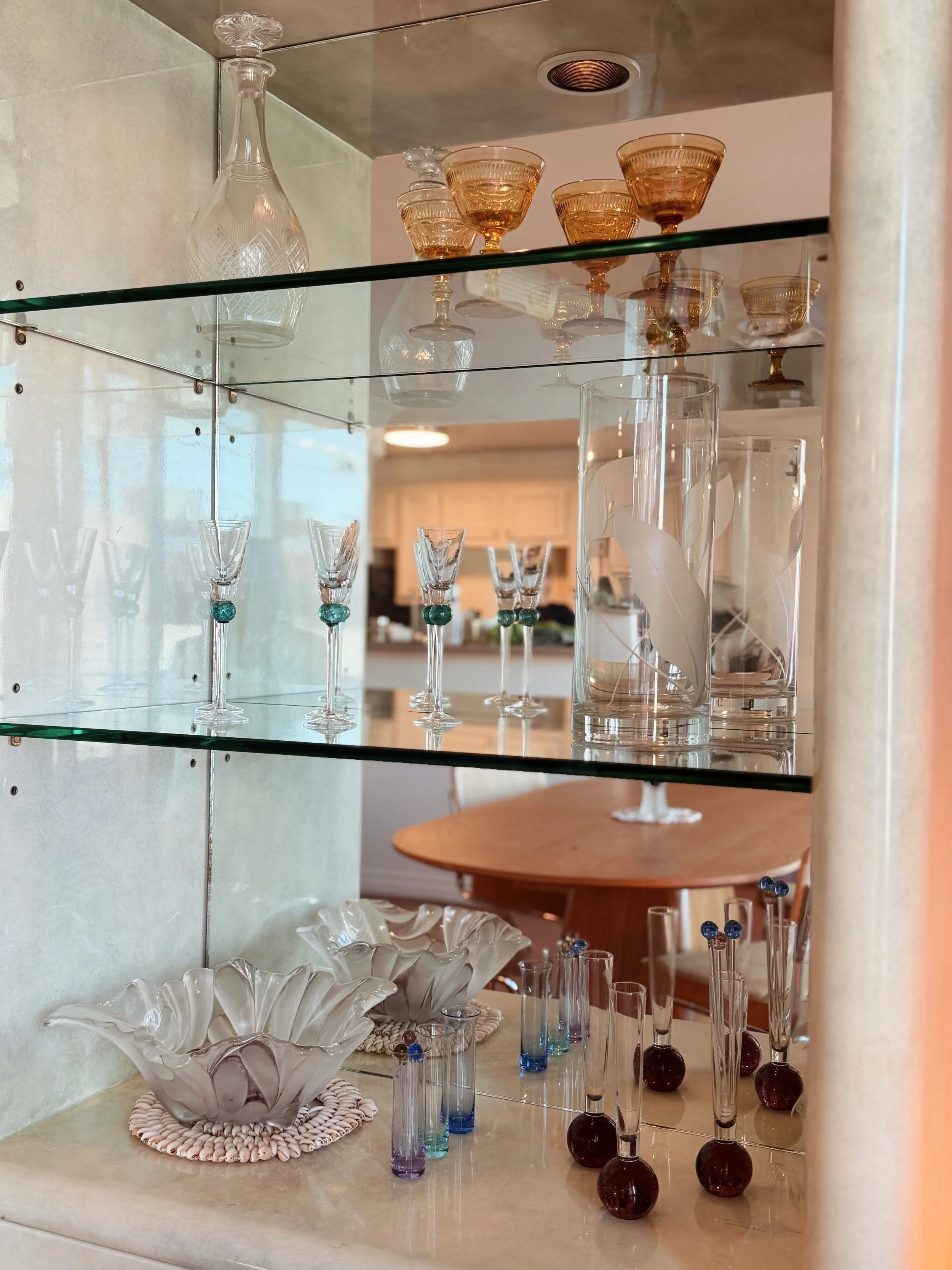 A display of various glassware including gold-rimmed glasses, blue-tinted vases, and a large decorative white glass bowl on a beige countertop with a mirror behind it.