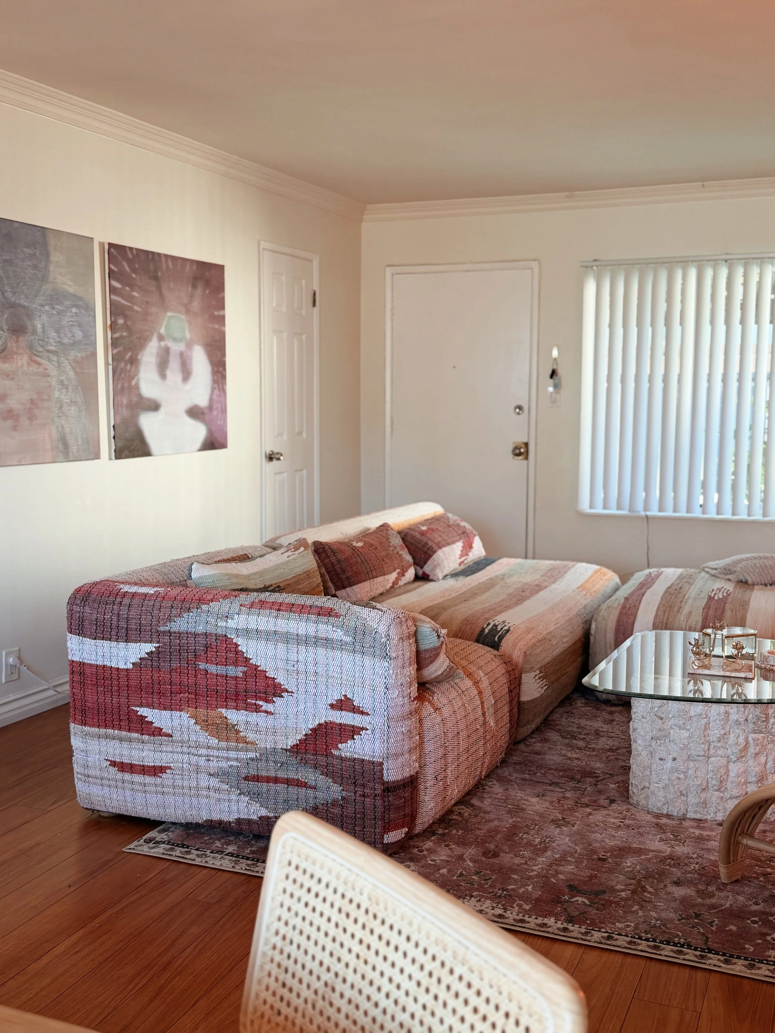 A cozy living room with a patterned couch, a glass coffee table, a rug, and artwork on the cream-colored wall near a door and window with vertical blinds.