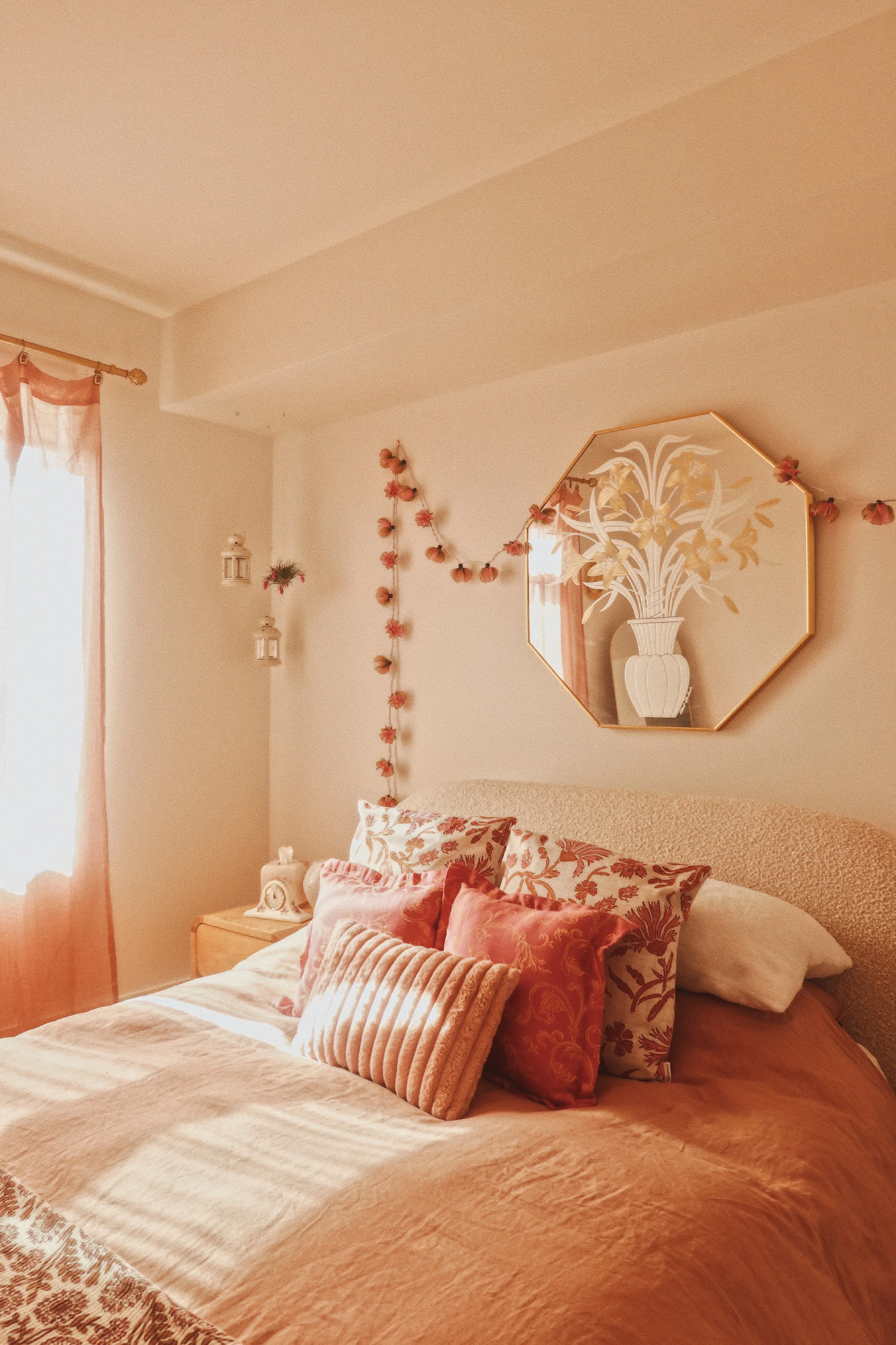 A cozy bedroom corner with a bed, multiple pillows in floral and striped patterns, a nightstand with a vintage alarm clock and a small decorative item, a wall mirror with a flower and vase design, and wall decorations including hanging peach-colored 