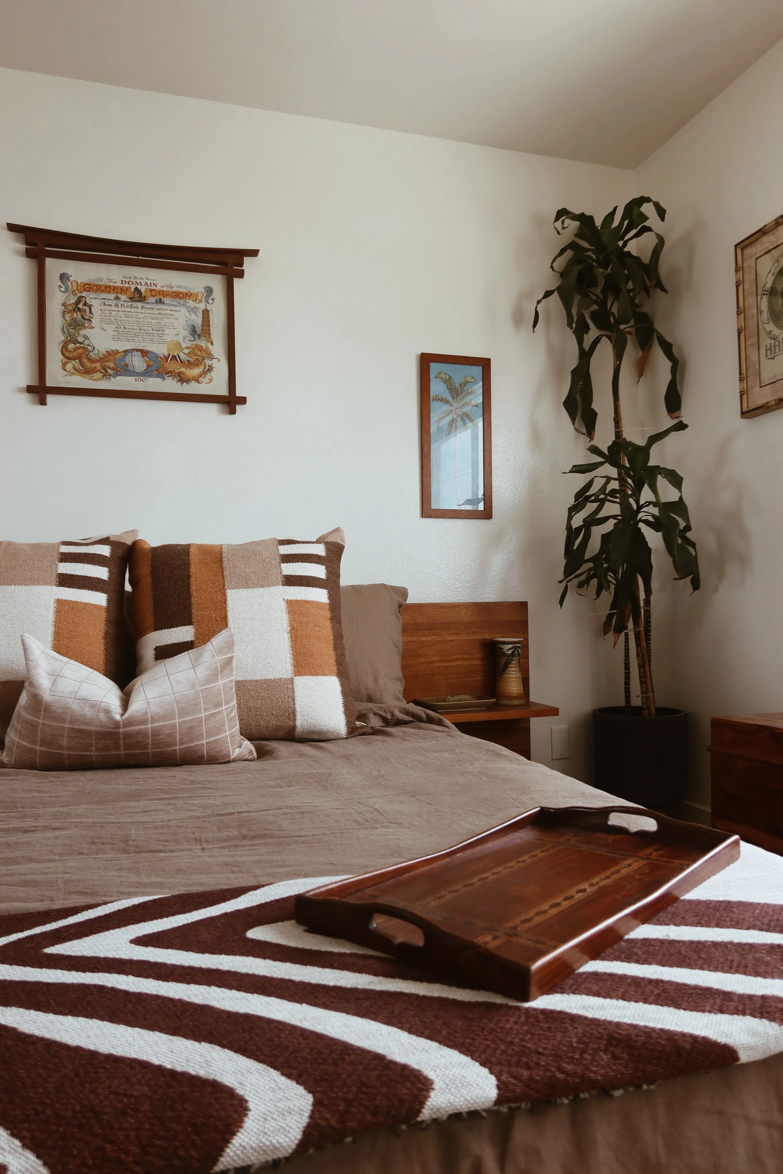 A bedroom corner with a bed, pillows, a wooden tray, a large potted plant, framed wall art, and a patterned rug.