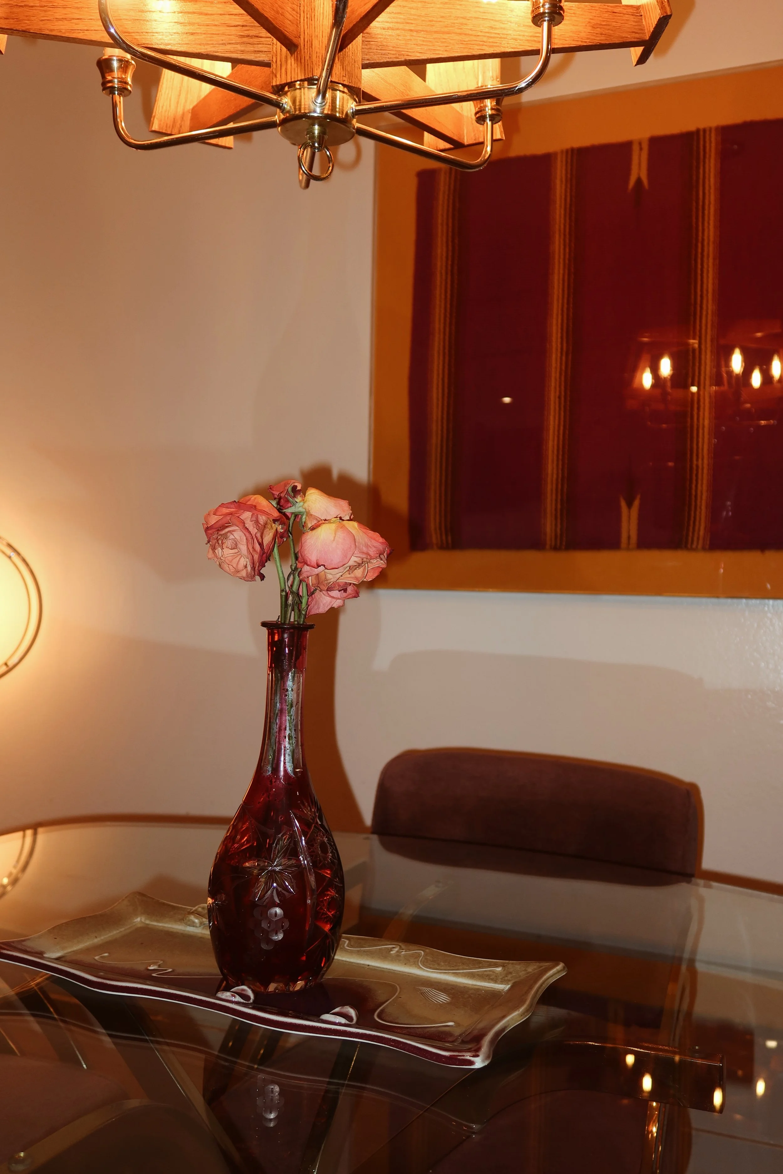 A dining room scene with a red glass vase holding pink flowers on a metallic tray, a wooden wall art piece, a brown upholstered chair, a glass table, a wall lamp, and a chandelier.