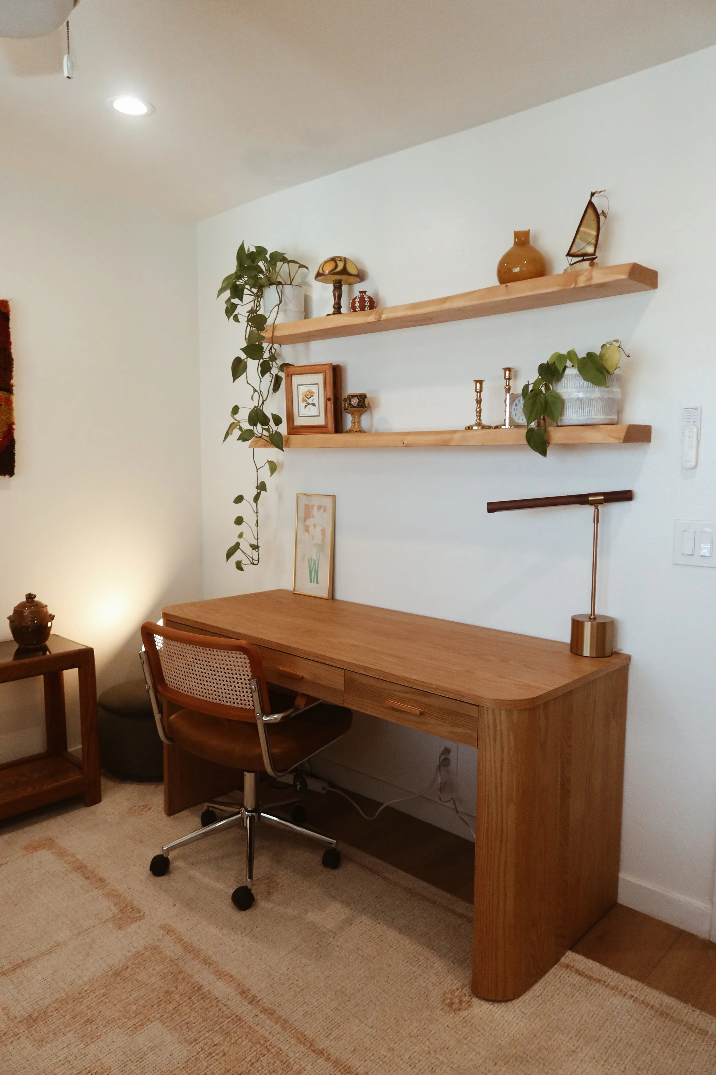 A home office with a wooden desk, an office chair, two wall-mounted shelves with decorative items, including plants, vases, a framed art piece, two candlesticks, a small boat model, and a lamp on the desk.