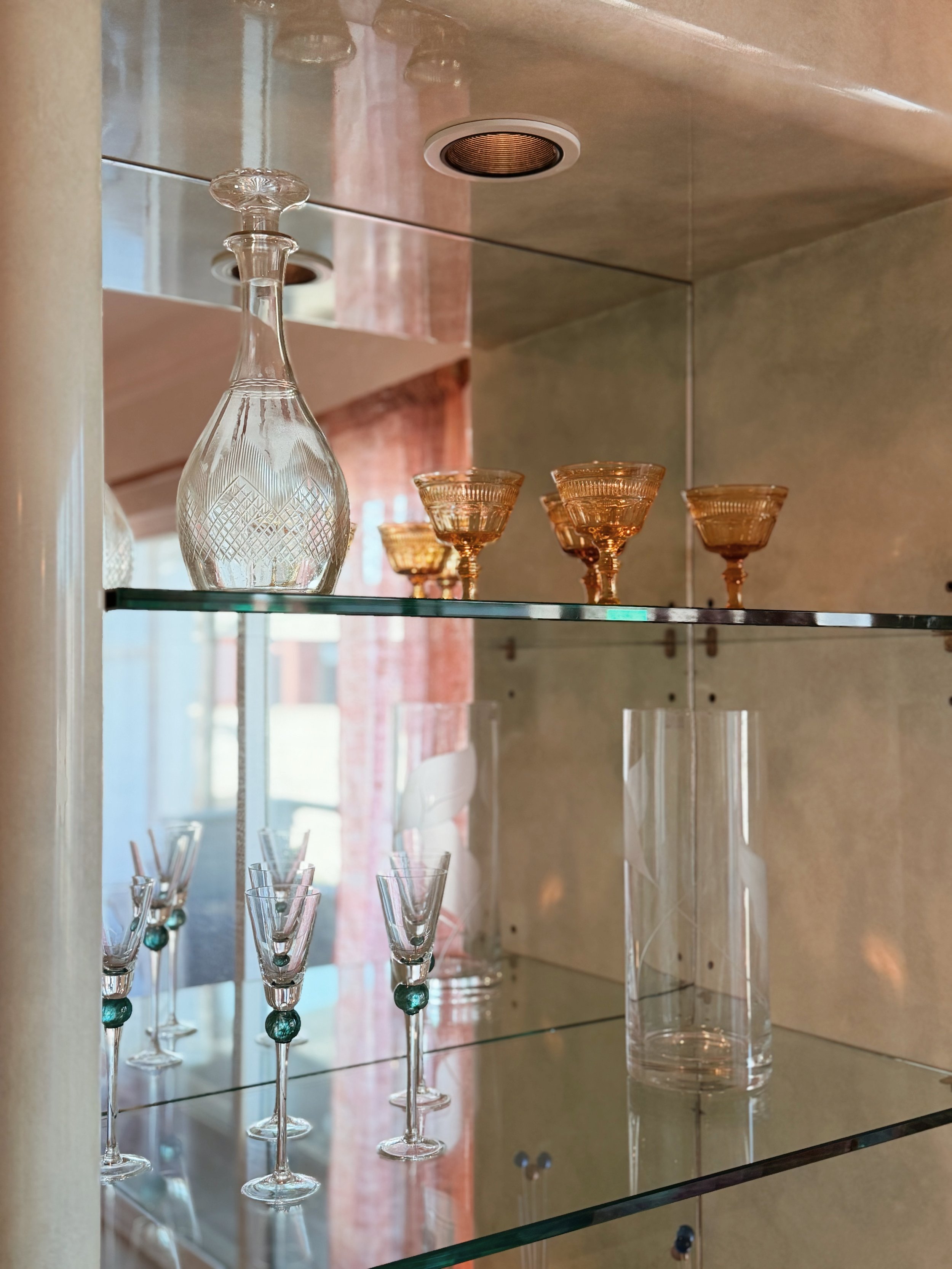 Display of various glassware including a large crystal decanter, pink glasses, and small glasses with green beads on a glass shelf, with a mirror reflecting the scene.