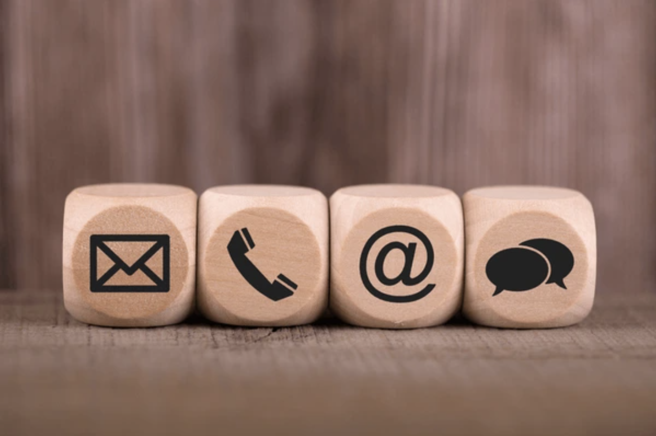 Four wooden blocks with black icons: email, phone, at symbol, and speech bubbles, on a wooden surface.
