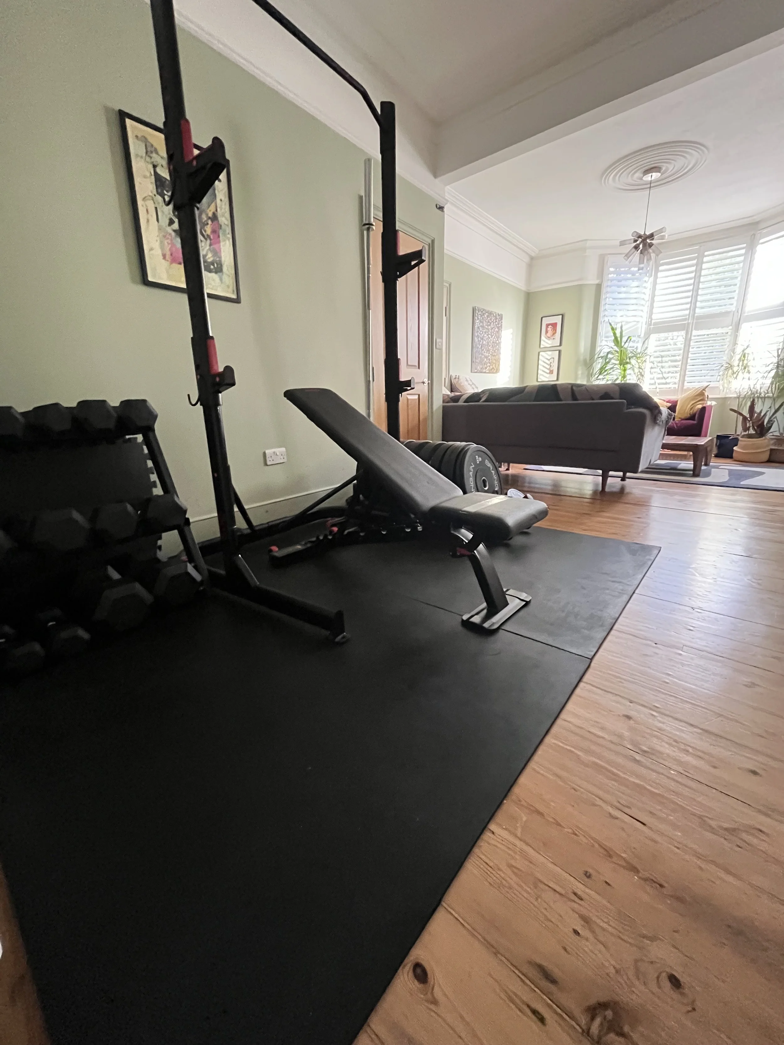 The home gym in Walthamstow where the personal training sessions can take place. Including black workout bench, dumbbells, and a squat rack on a black mat, with a living room with a sofa, plants, and large windows in the background.