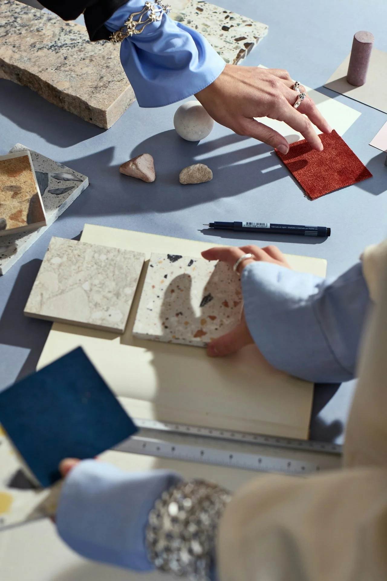 Person selecting fabric swatch among samples of stone and tile samples on a table, with a notebook and pen nearby.