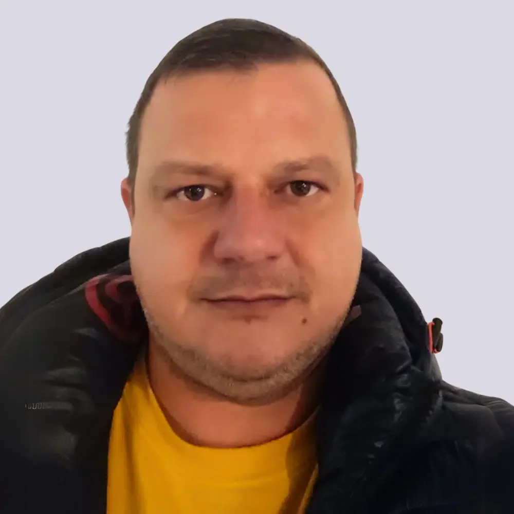 Nahaufnahme eines Mannes mit kurzen dunkelbraunen Haaren, braunen Augen, schwarz-weißem Bart und leichter Gesichtsbehaarung, trägt eine schwarze Jacke mit rotem Reißverschluss und ein gelbes T-Shirt, vor hellem Hintergrund.