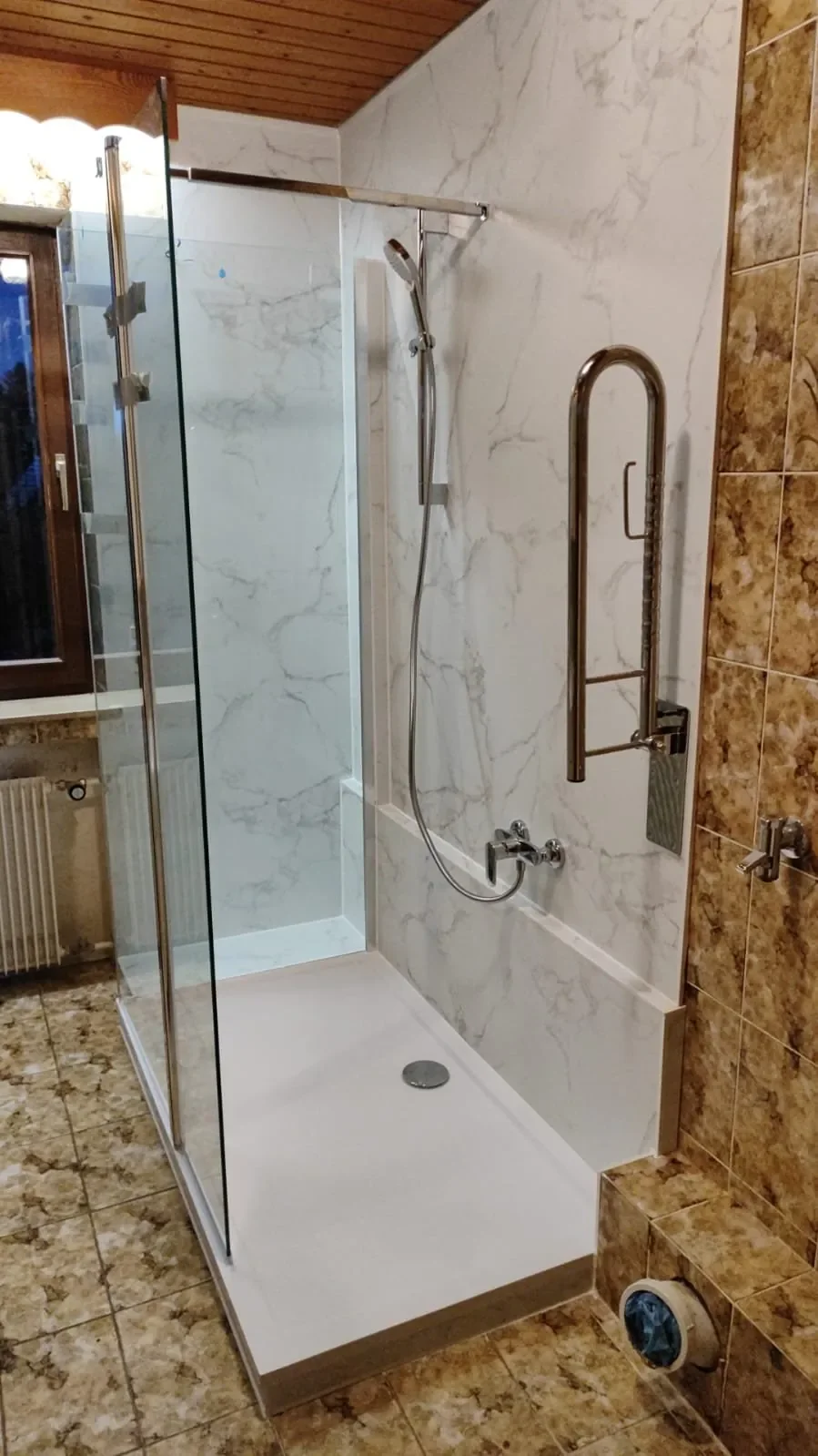 Moderner barrierefreier Duschbereich mit schwenkbarem Duschkopf, Edelstahlgriffen und Glastür in einem Badezimmer mit Marmorfliesen.