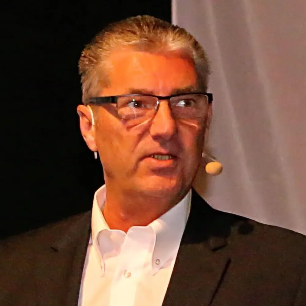 Ein Mann mit grauem, kurzen Haar, trägt eine schwarze Brille, eine weiße Krawatte und ein schwarzes Jackett, spricht bei einer Veranstaltung.