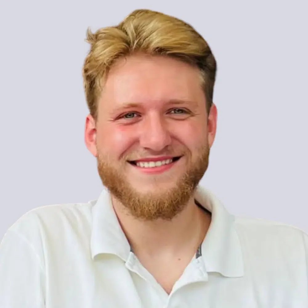 Ein lächelnder Mann mit blonden Haaren, Bart und weißem Hemd vor einem blauen Hintergrund.