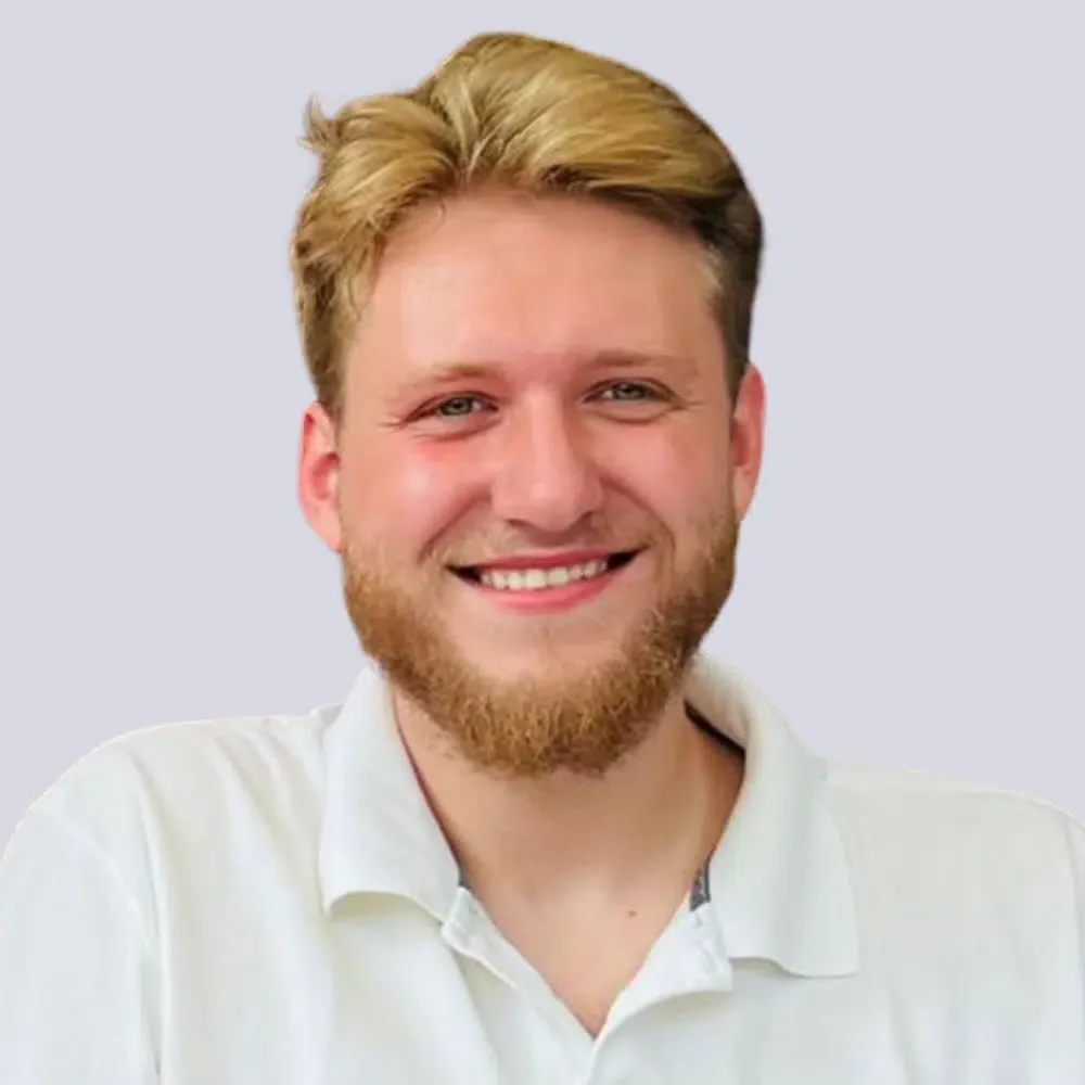 Ein lächelnder Mann mit blonden Haaren, Bart und weißem Hemd vor einem blauen Hintergrund.