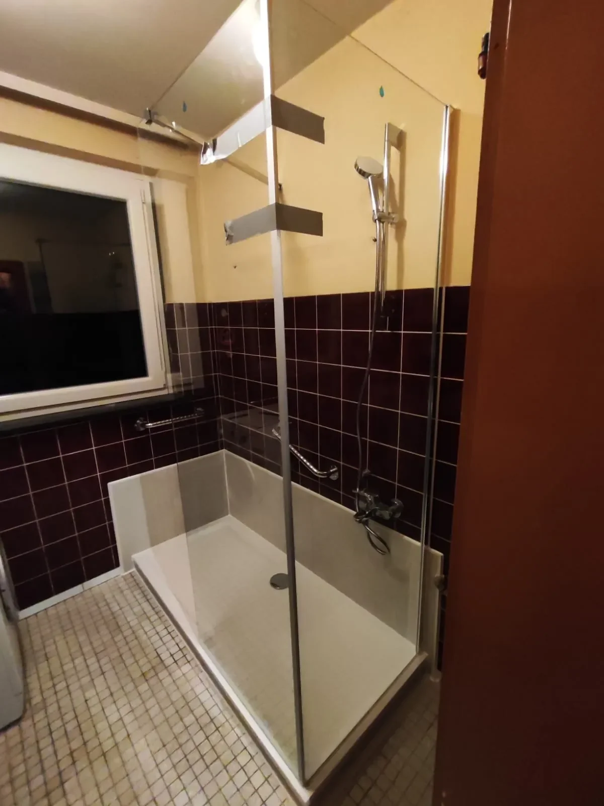 Kleines Badezimmer mit Dusche, dunklen Fliesen, einer Fensteröffnung und gelber Wandfarbe.