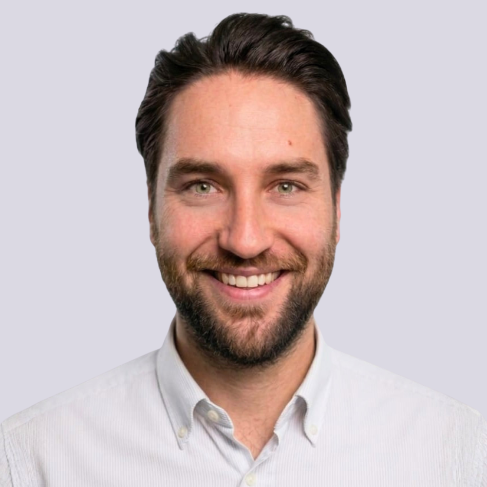 Ein junger Mann mit kurzen braunen Haaren trägt ein weißes Hemd, lächelnd vor hellem Hintergrund.