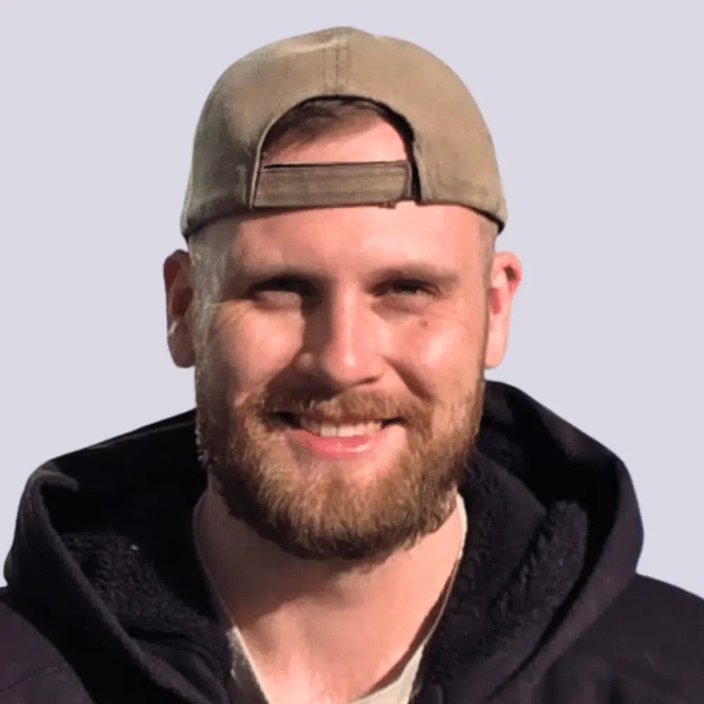 Ein Mann mit Bart trägt einen schwarzen Hoodie und eine beigefarbene Cap, lächelt und schaut in die Kamera.