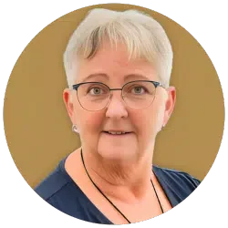 Portrait einer älteren Frau mit kurzen, grauen Haaren und Brille, lächelnd vor beigem Hintergrund.