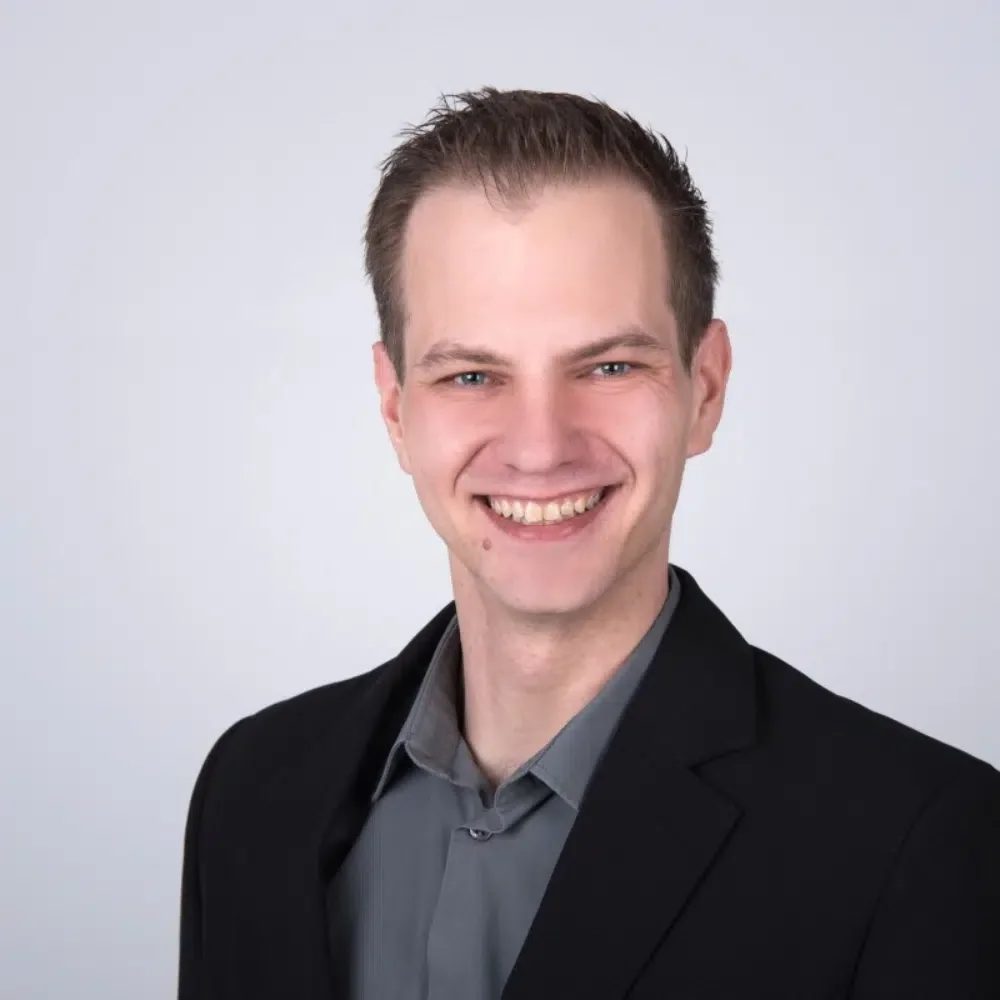 Ein junger Mann mit kurzen braunen Haaren trägt einen schwarzen Anzug und ein graues Hemd, lächelnd vor hellem Hintergrund.