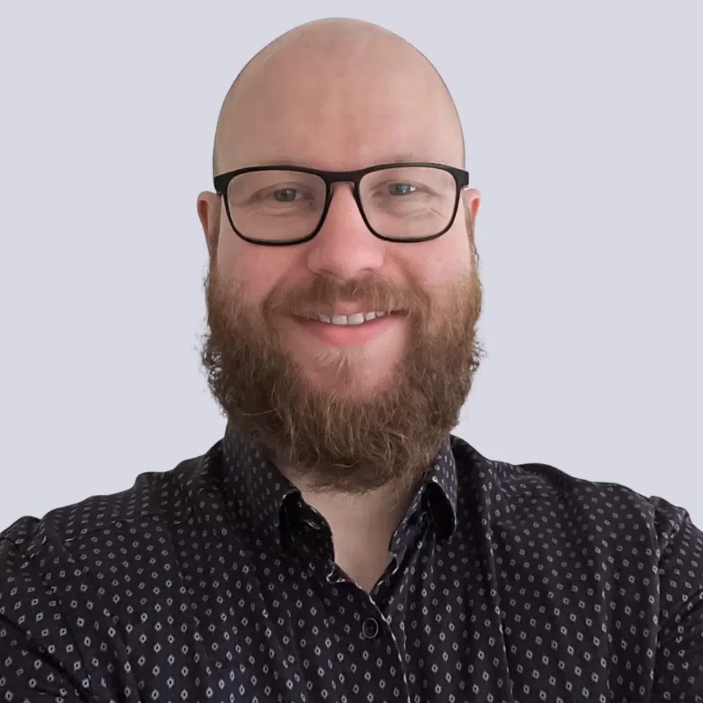 Ein lächelnder Mann mit Brille und Bart vor einem hellen Hintergrund.