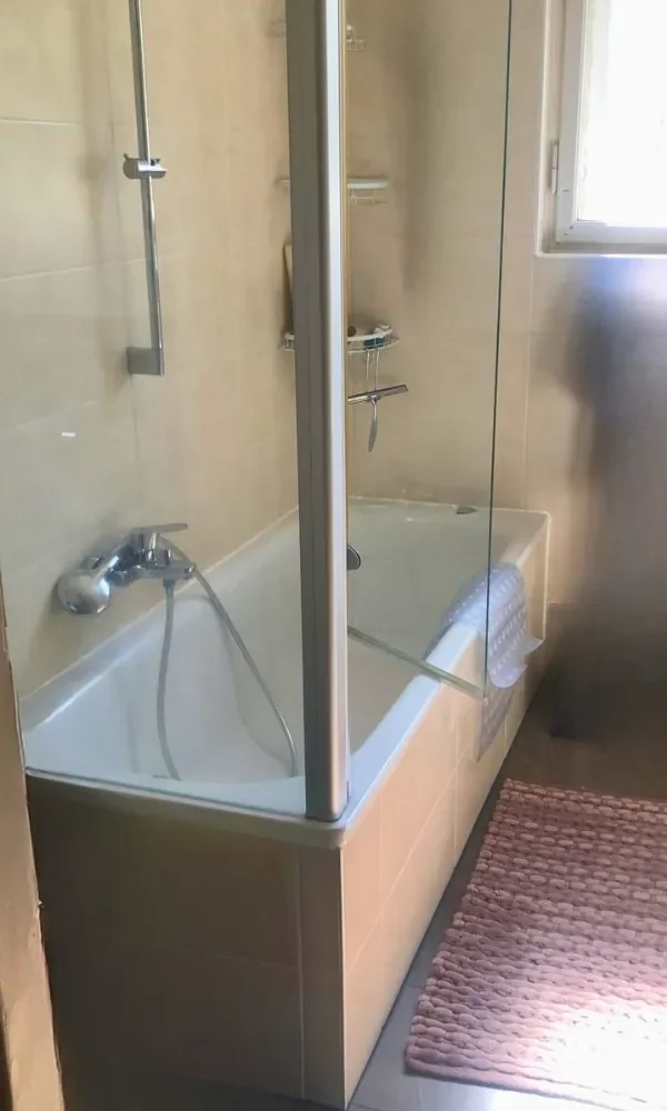 Ein Badezimmer mit Badewanne, Duschwand, Wasserhahn und Duscharmatur. Es gibt eine Fensterseite und einen Badezimmerteppich auf dem Boden.