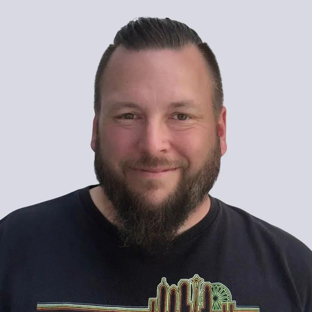 Ein Mann mit dunklem Haar, Bart und Kinnbart, trägt ein schwarzes T-Shirt mit einem bunten Skyline-Design, lächelt vor hellem, neutralem Hintergrund.