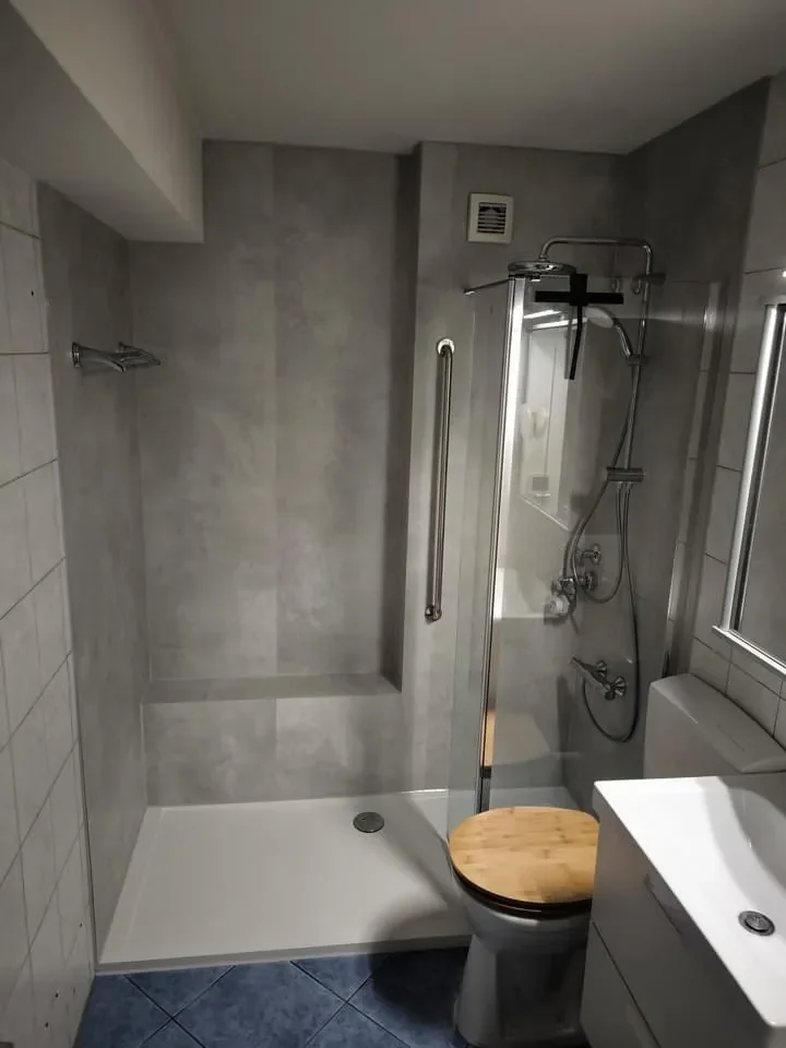 Ein kleines Badezimmer mit einer Dusche, einem WC mit Holzsitz, einem Waschtisch und einem Fenster, die Wände sind grau gefliest.