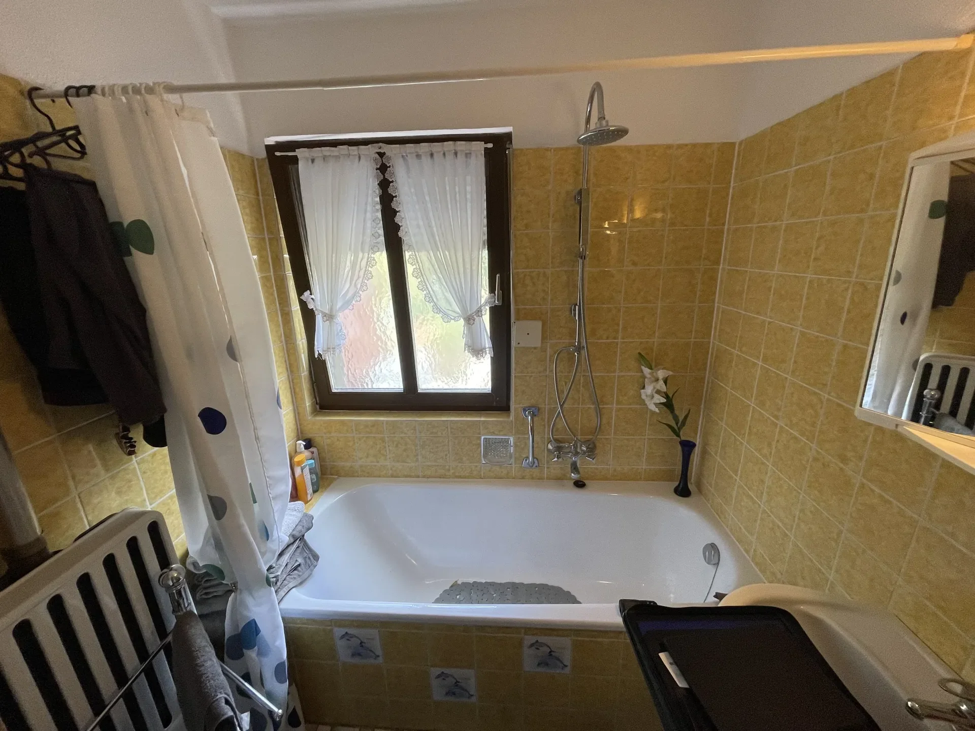 Ein Badezimmer mit gelben Fliesen, einem Fenster mit weißen Vorhängen, einer Dusche mit Regenduschkopf, einem Spiegel und einem Waschbecken. Es sind verschiedene Hygieneprodukte auf einem Regal neben der Badewanne und ein Handtuch an einem Heizkörper. Ein kleiner Beistelltisch mit einem schwarzen Gegenstand ist sichtbar.