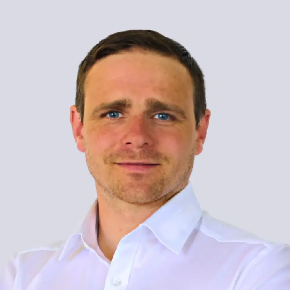 Der Mann trägt ein weißes Hemd und hat braune Haare, blaue Augen, leicht rasierten Bart. Der Hintergrund ist hell grau.