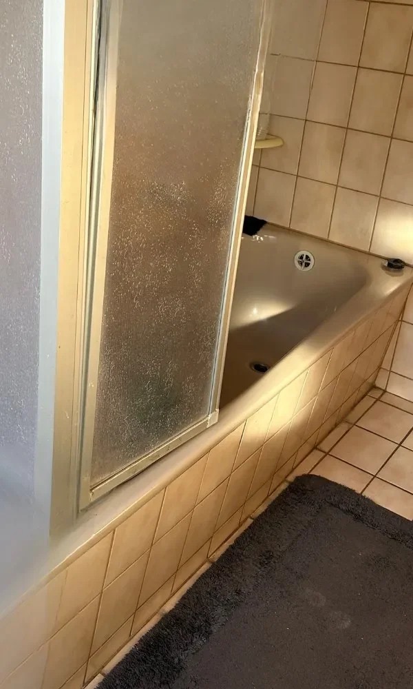 Badezimmer mit Badewanne, Duschtür, Fliesen und schwarzem Badvorleger.