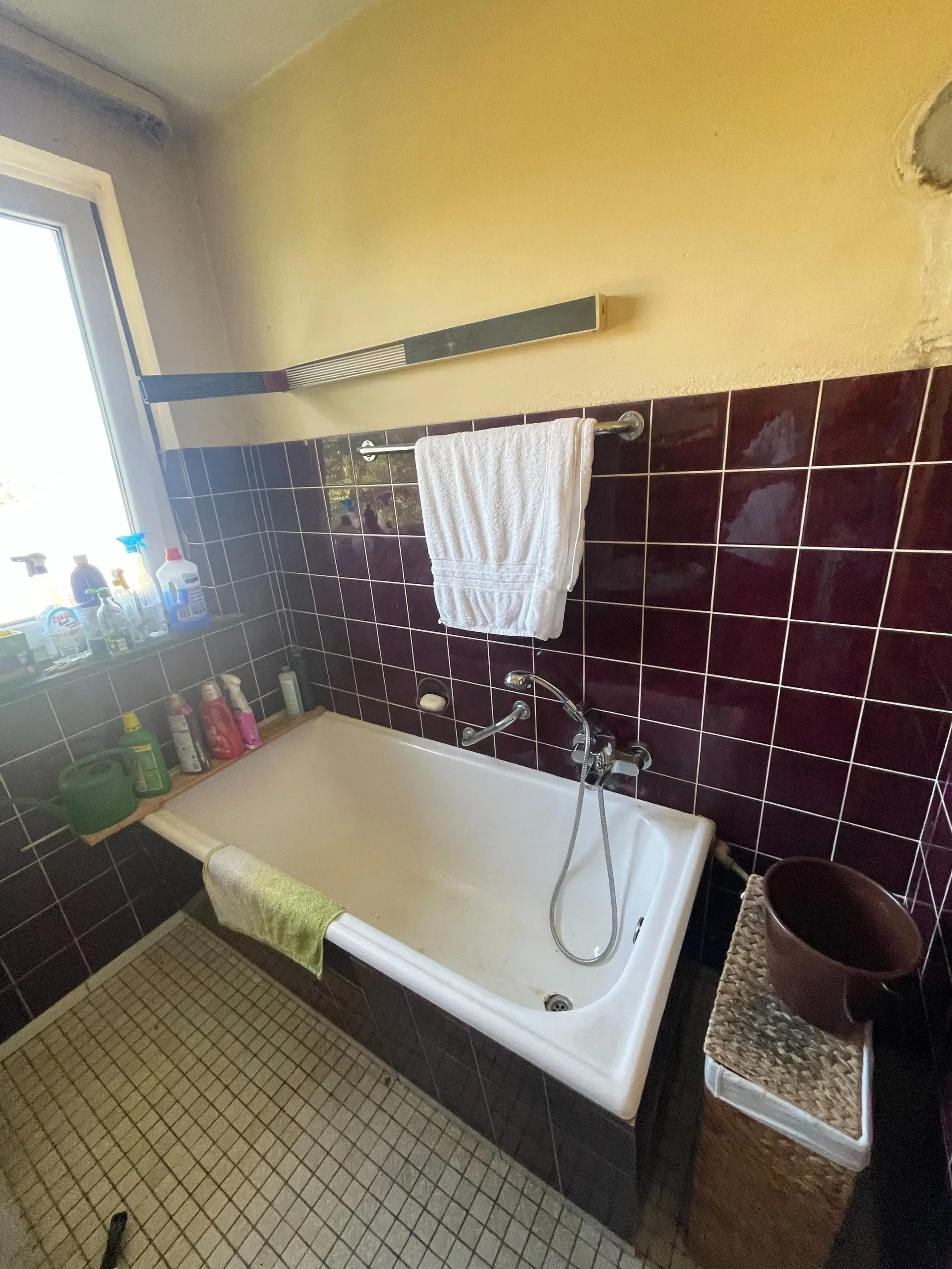 Ein Badezimmer mit braunen Fliesen, eine Badewanne, eine weiße Handtuchstange mit einem weißen Handtuch, eine Fensteröffnung mit Reinigungsmitteln und ein Korb mit Körben.
