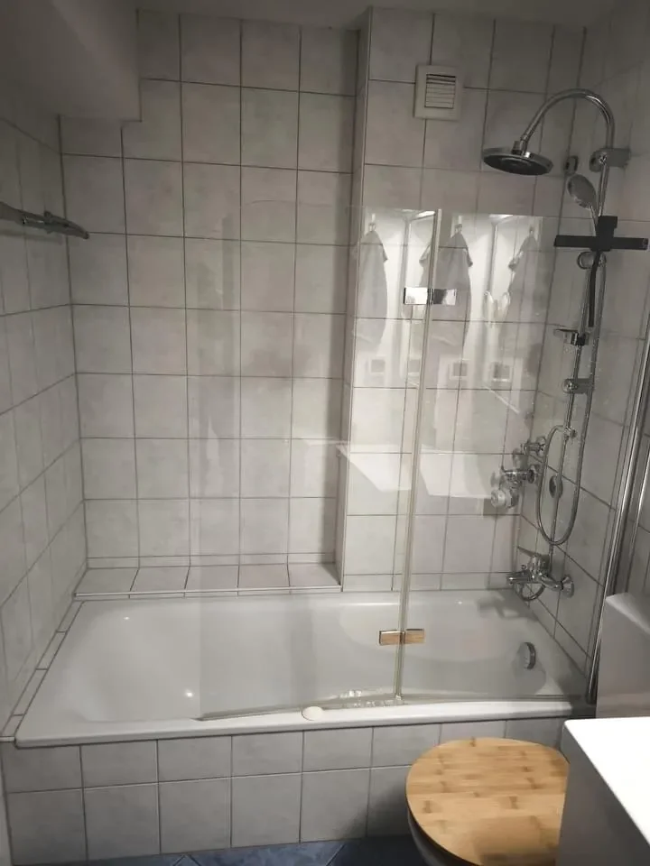 Badezimmer mit Duschbadewanne, Wandfliesen, Handbrause, Ventilator an der Wand, Holzdeckel auf einem Gegenstand im Vordergrund.