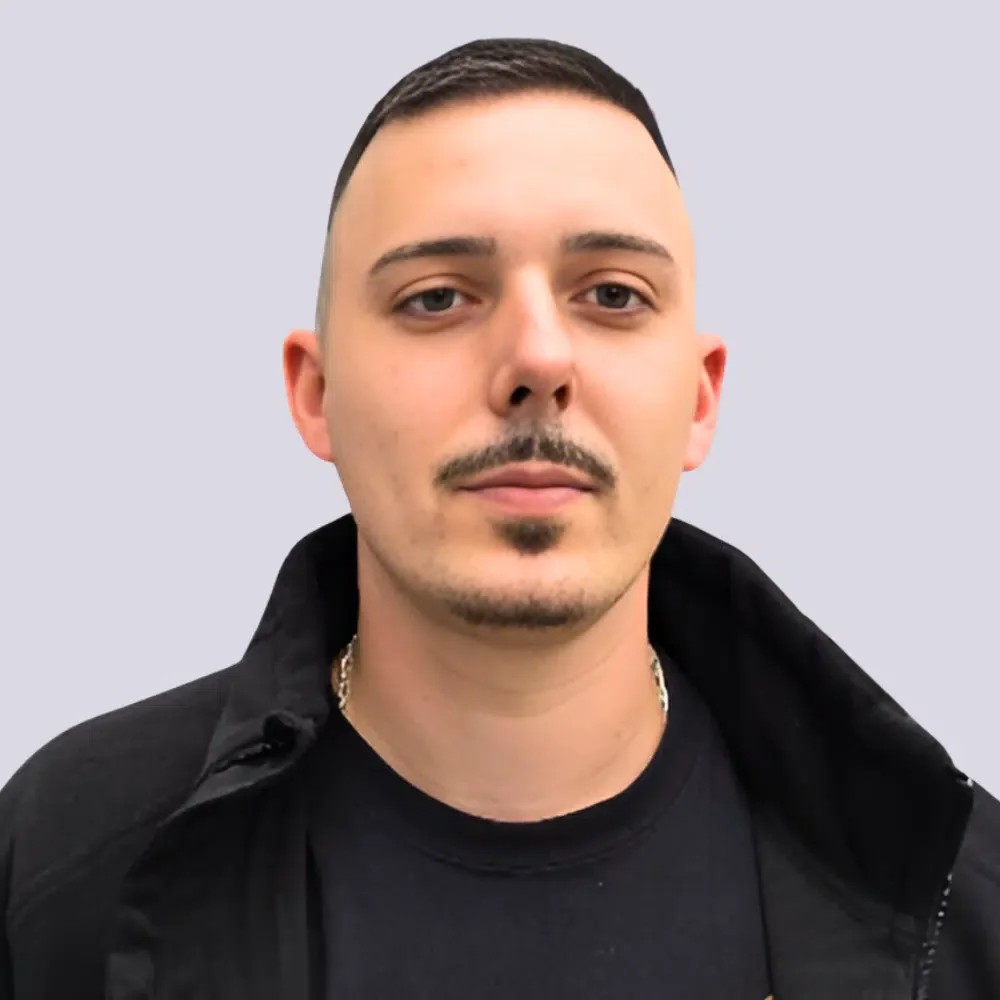 Porträt eines Mannes mit dunklem Haar, kurz geschnitten, und einem kleinen Bart, trägt dunkle Kleidung vor einem hellgrauen Hintergrund.
