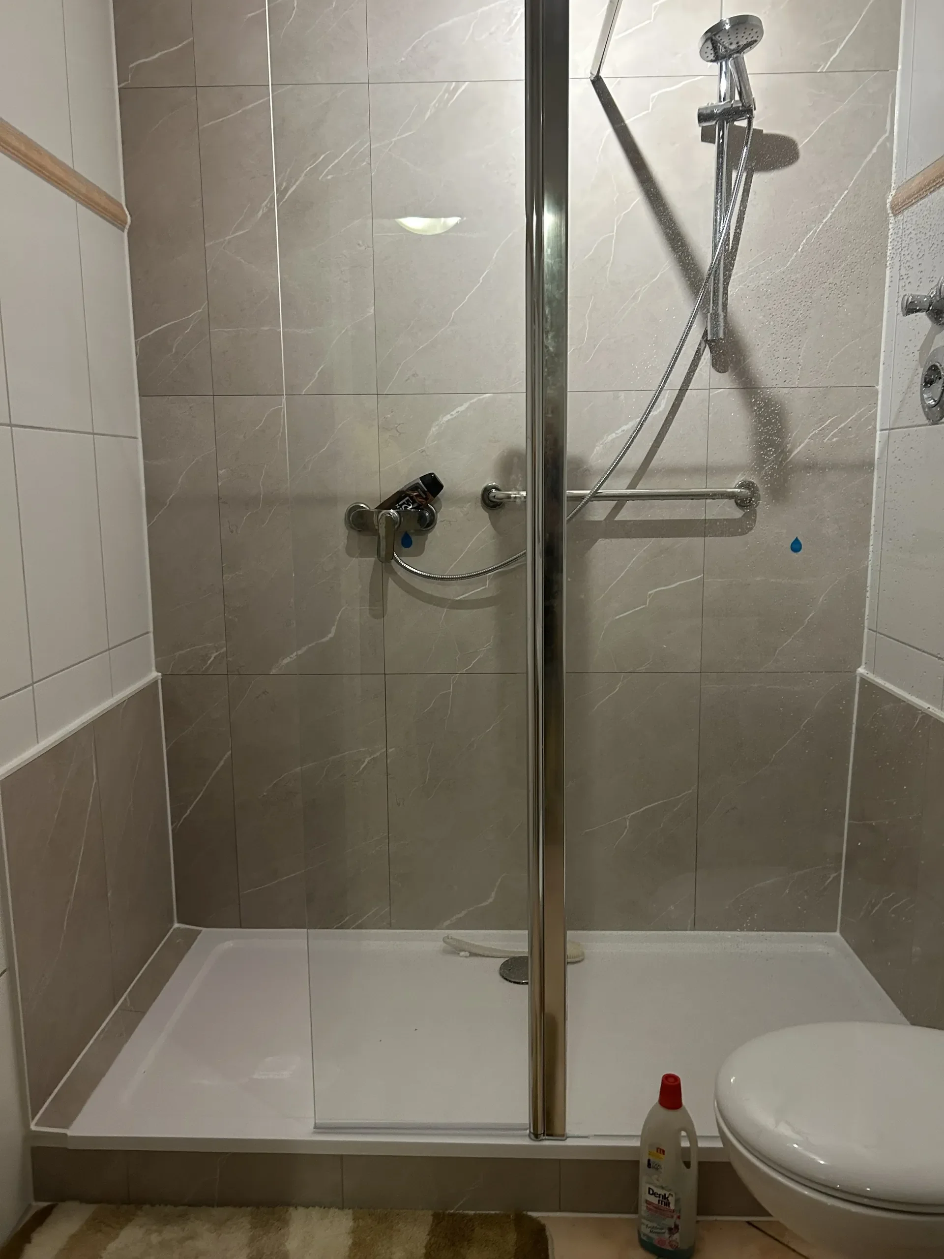 Ein Badezimmer mit einer Dusche, einer Toilette, einem Bodenfit und einer Flasche Reinigungsmittel vor der Toilette.