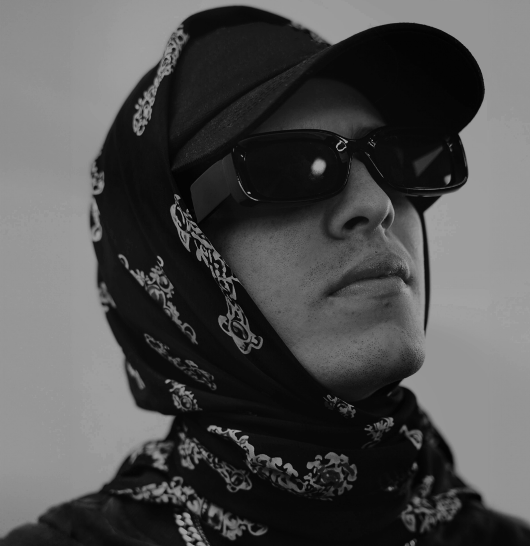 Retrato en blanco y negro de una persona con gafas oscuras, gorra y pañuelo en la cabeza, mirando hacia la izquierda.