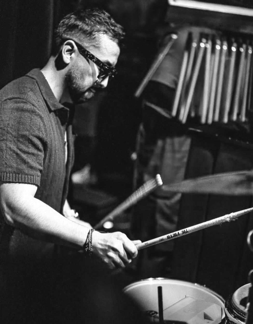 Hombre tocando la batería en un entorno oscuro, usando gafas y con barras de percusión y partituras en el fondo.