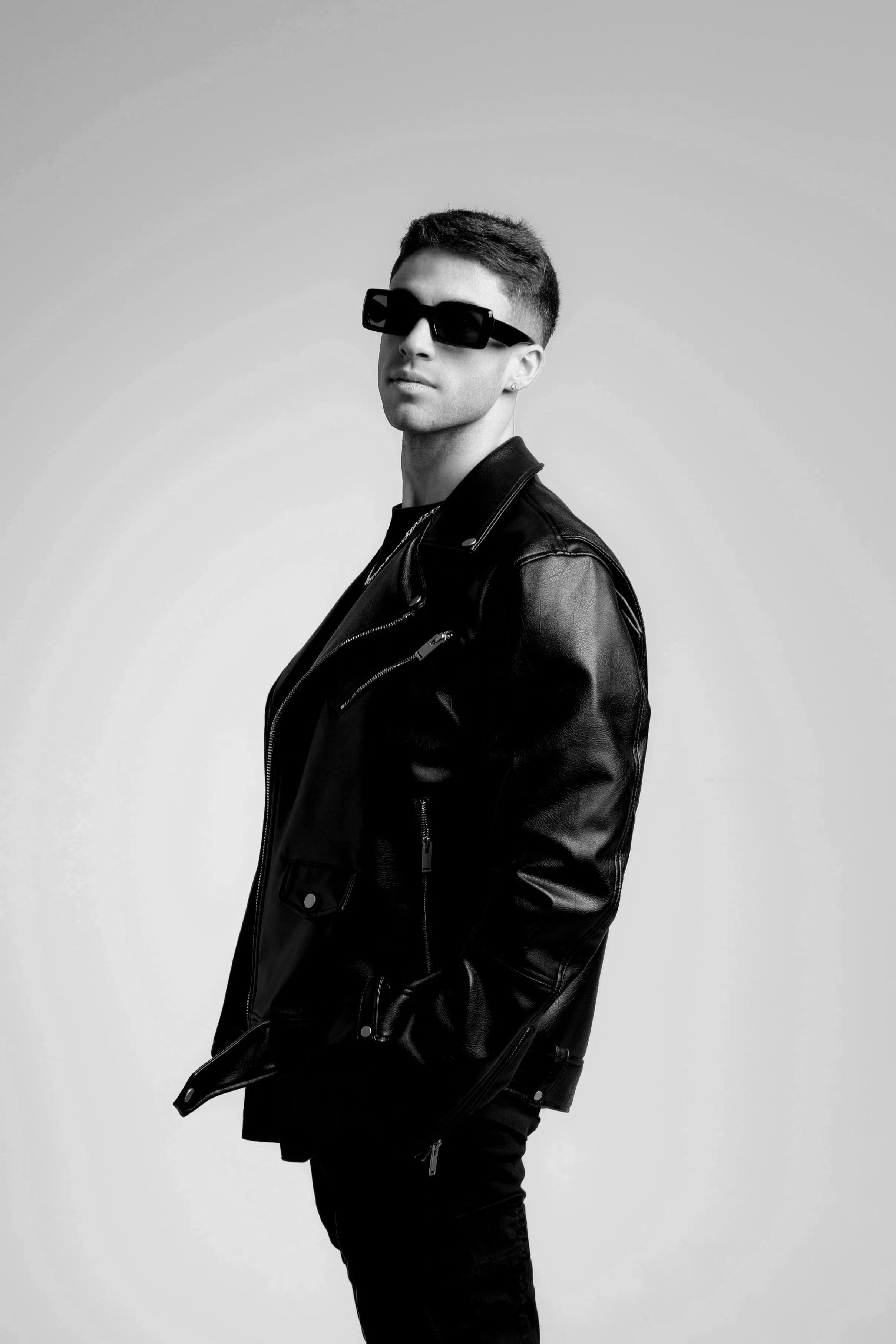 Joven con gafas de sol, chaqueta de cuero y pantalones negros posando en fondo gris.