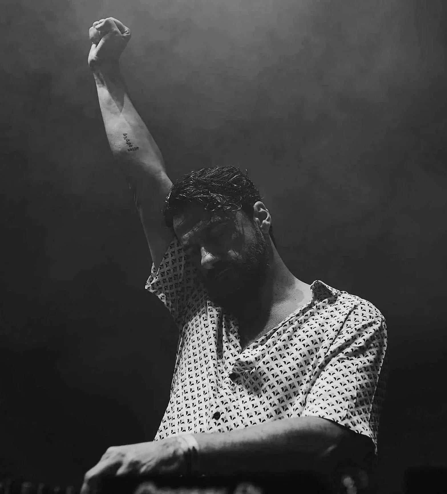 Hombre con barba y gafas de sol, levantando un brazo en un entorno oscuro, usando una camisa de patrón geométrico