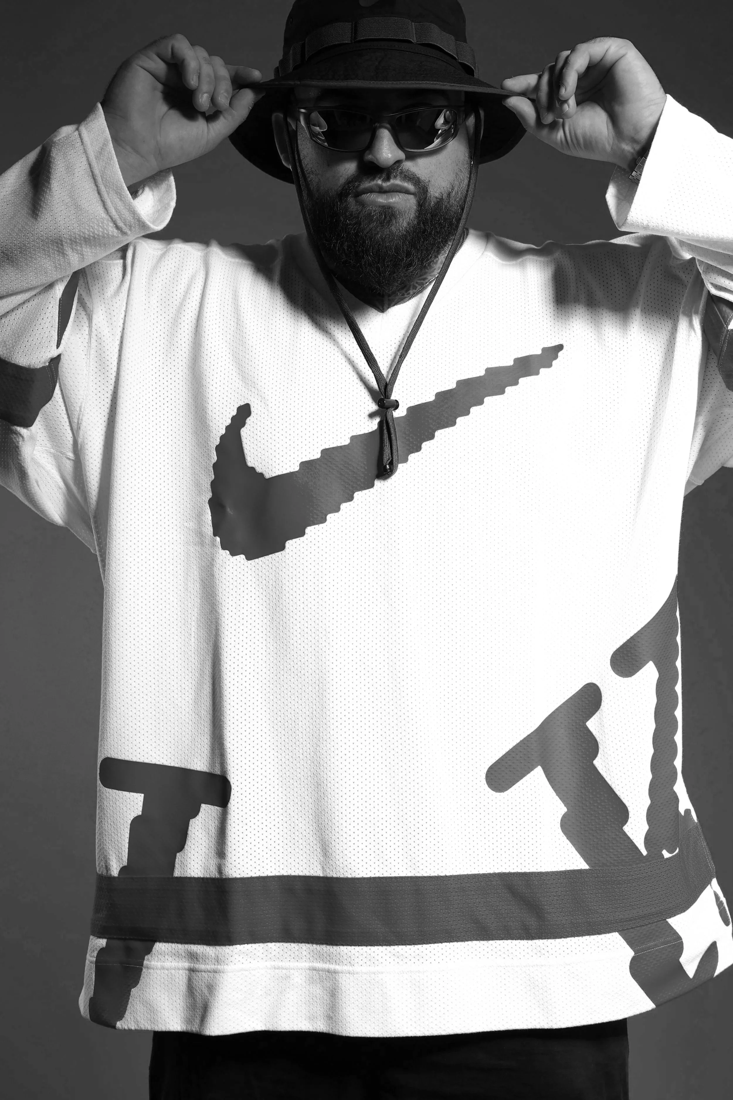 Hombre con sombrero, gafas de sol y barba, vistiendo camiseta blanca con logotipo de Nike en la parte frontal, en un fondo gris.