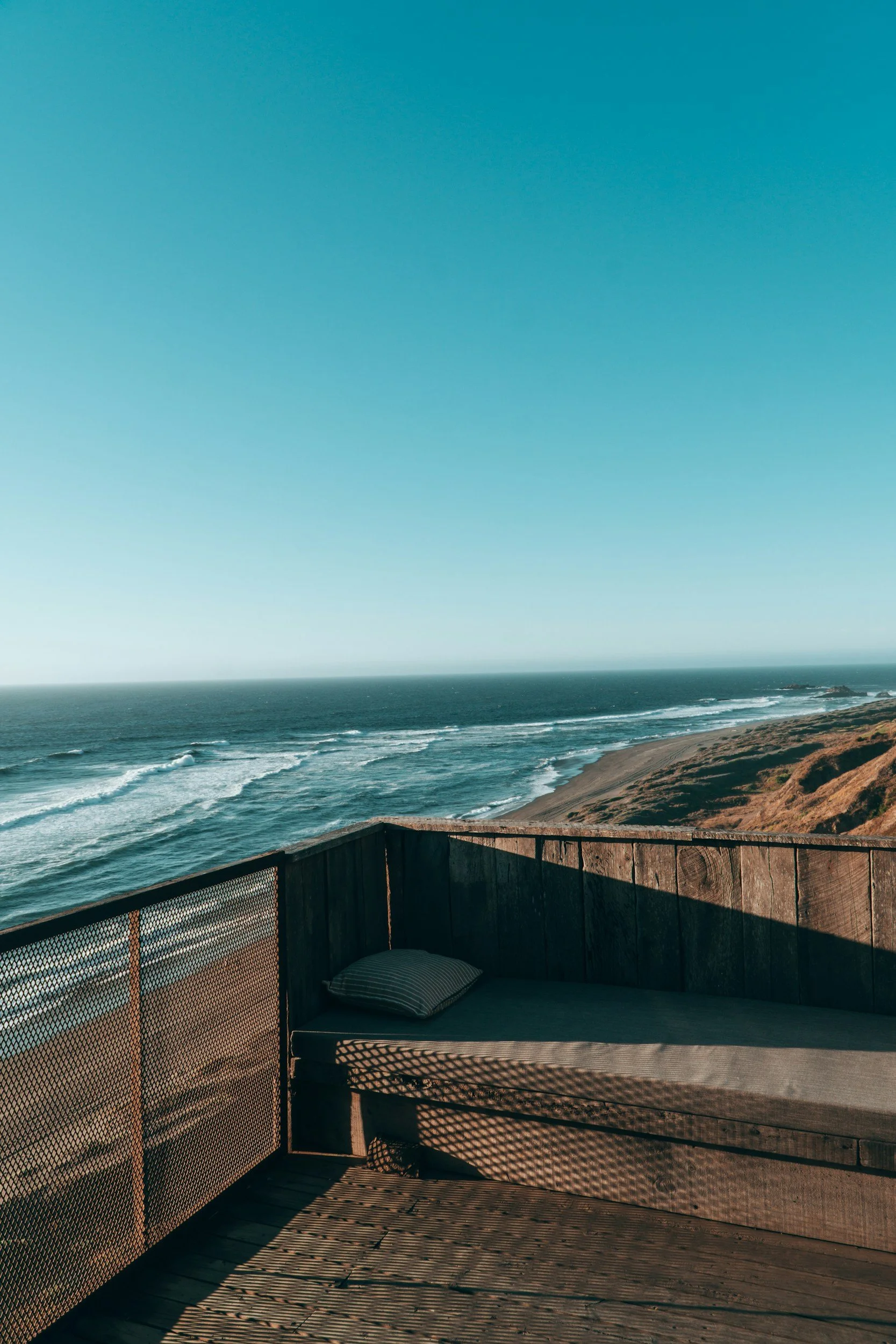 An outdoor wooden deck with a cushioned lounge area overlooking the ocean, on a sunny day with clear blue sky.