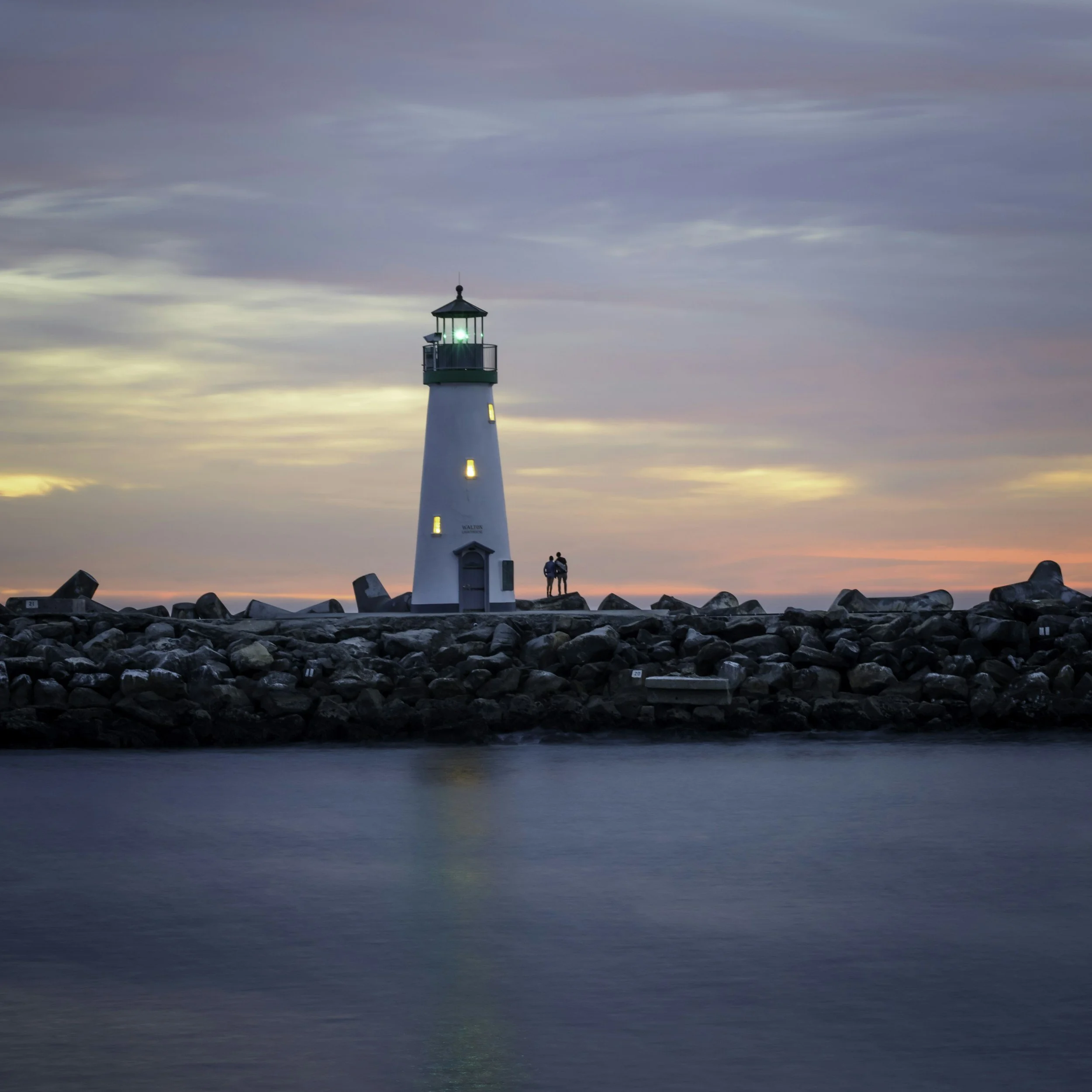 A lighthouse on a rocky pier at sunset with a pink and purple sky, calm water in the foreground, and two people standing near the lighthouse.