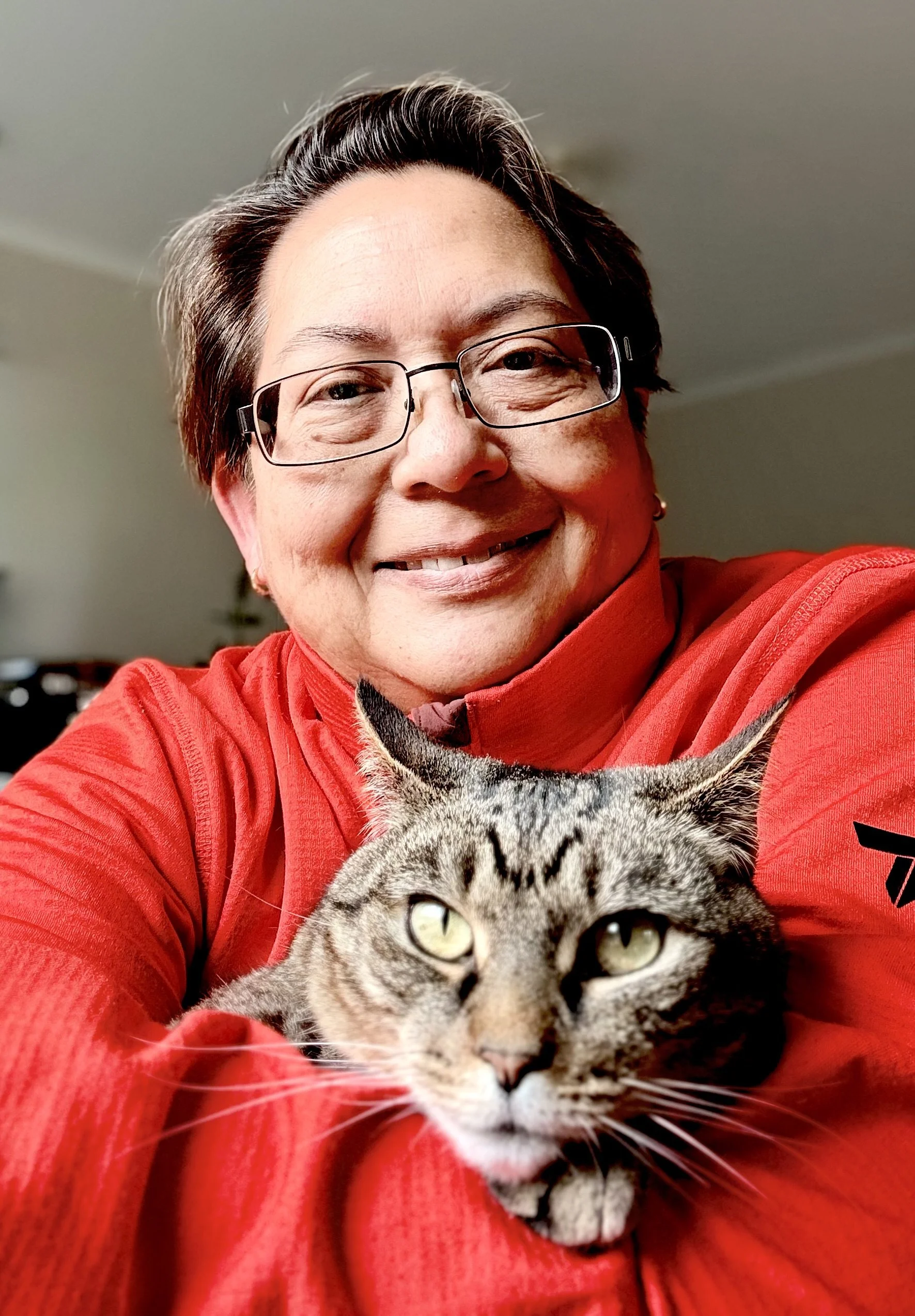 A woman smiling and taking a selfie with a tabby cat resting on her arm, indoors.
