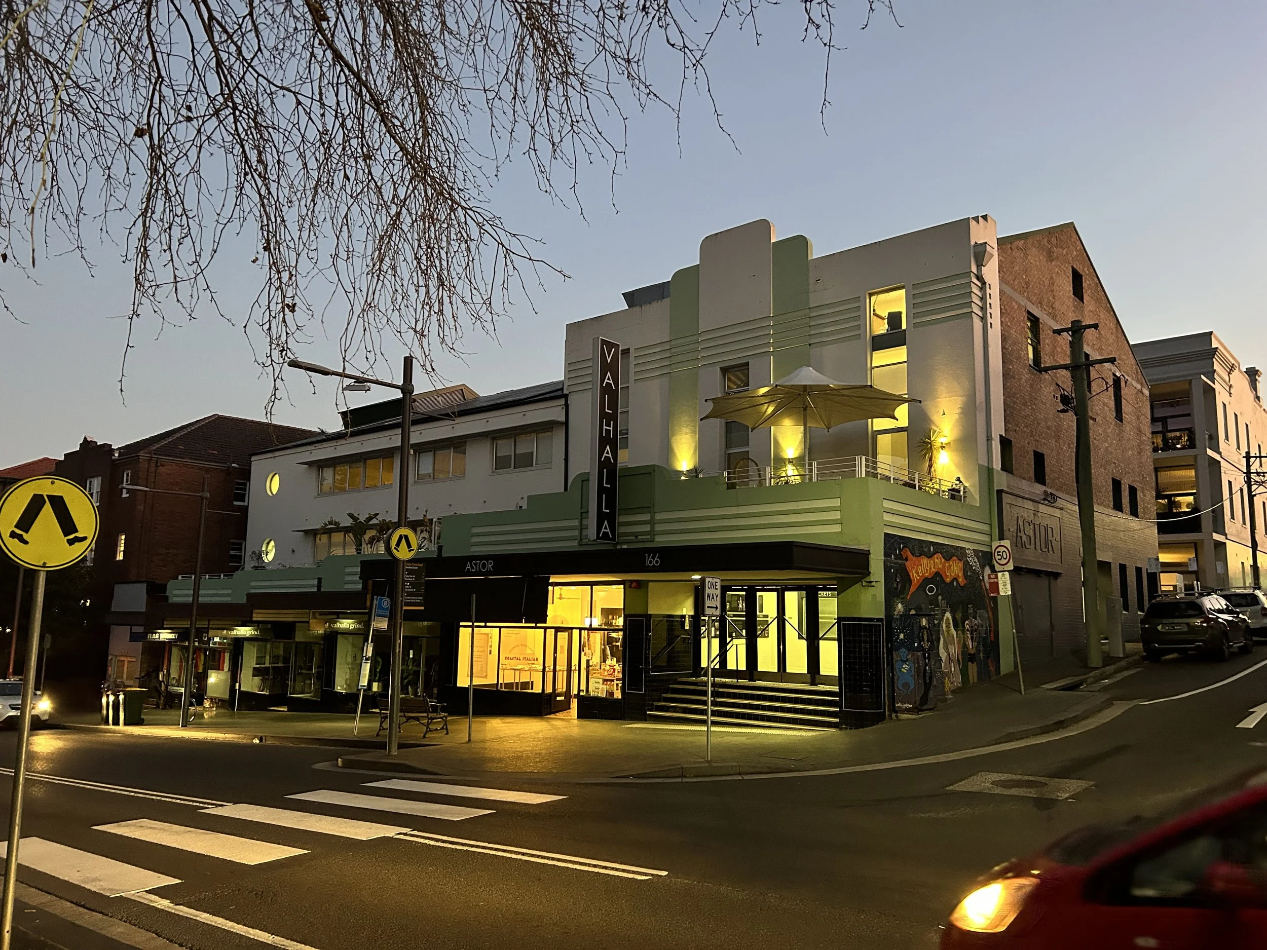 Art Deco style building called Valhalla with art murals on the side, illuminated with warm yellow lighting, street view with pedestrian crosswalk and street signs at dusk
