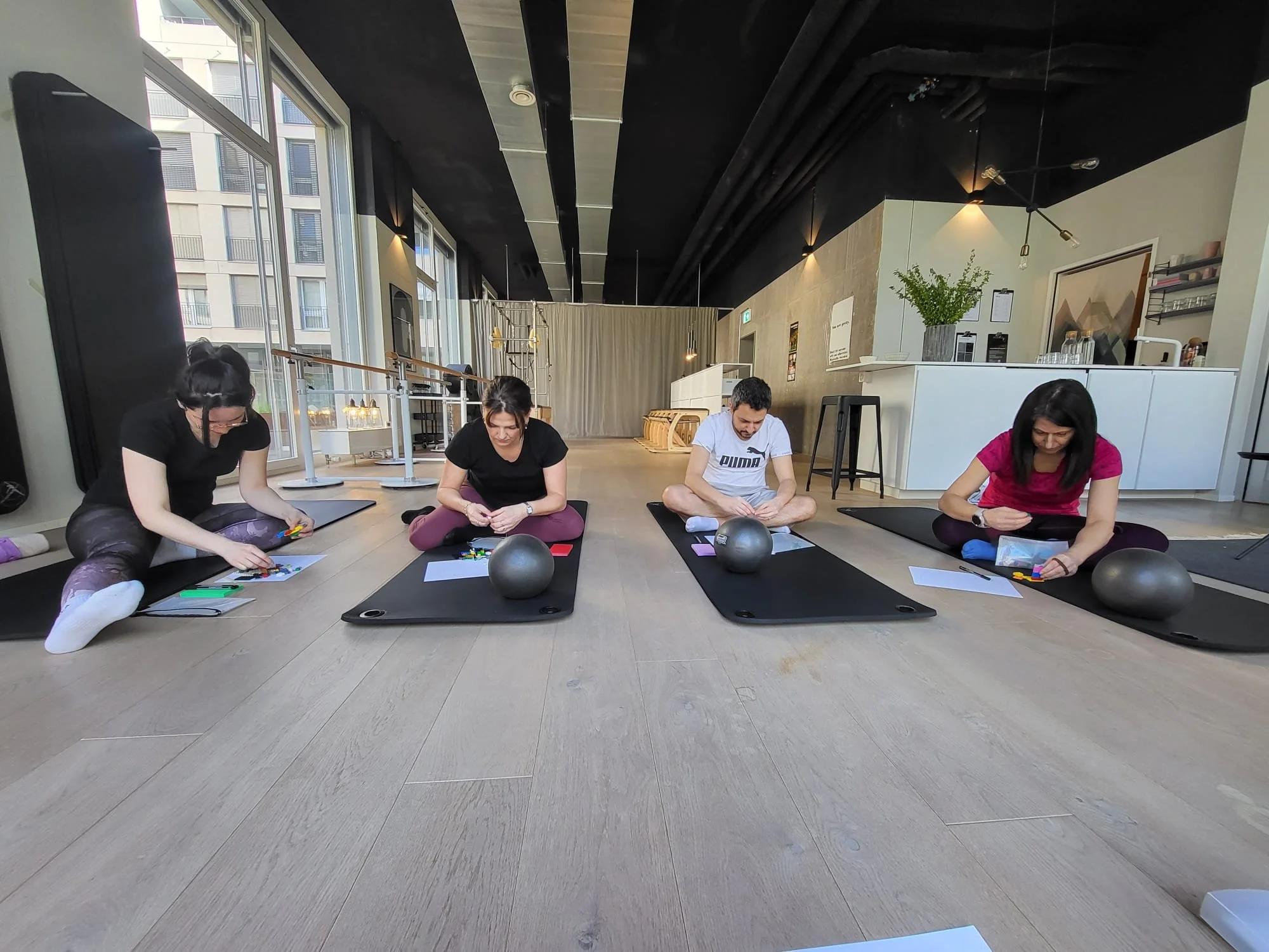 Four individuals sitting on yoga mats, engaged in a craft activity inside a modern room with large windows and natural light.