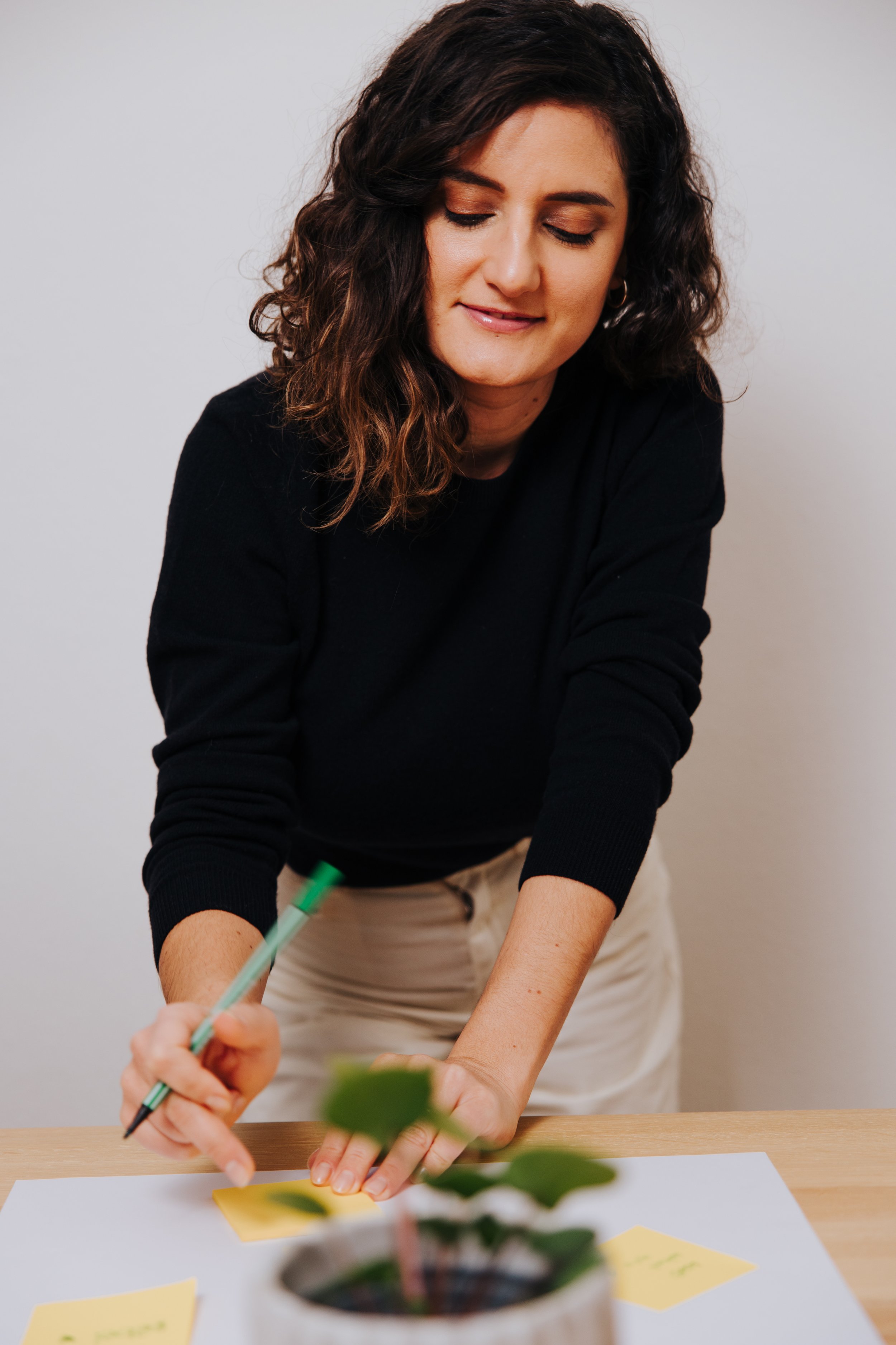A woman with curly brown hair wearing a black sweater and beige pants is standing at a desk, writing on yellow sticky notes with a green pen.