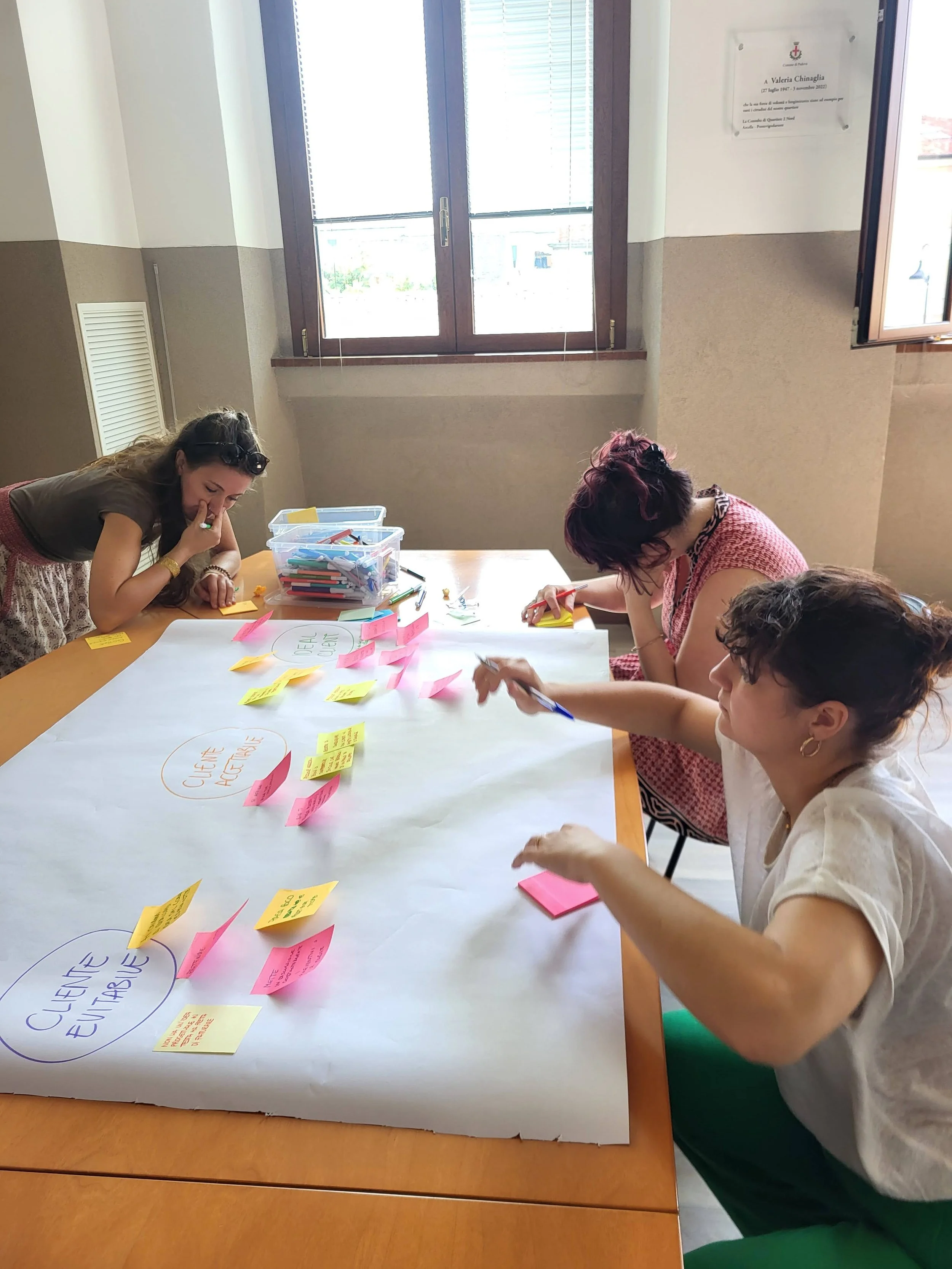 Three women working together on a project at a table with colorful sticky notes and markers in a well-lit room with large windows.